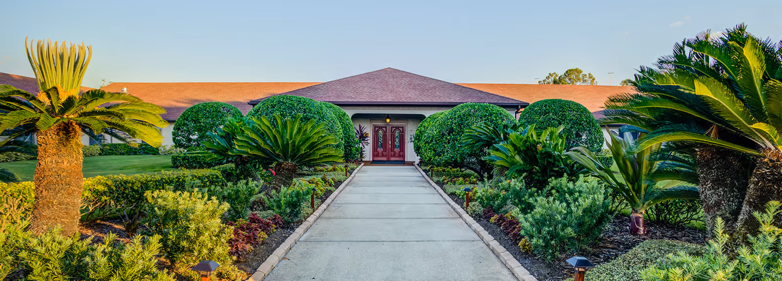 Walkway leading to the front double doors of a low-rise building, bordered by neatly trimmed shrubs and tropical plants.