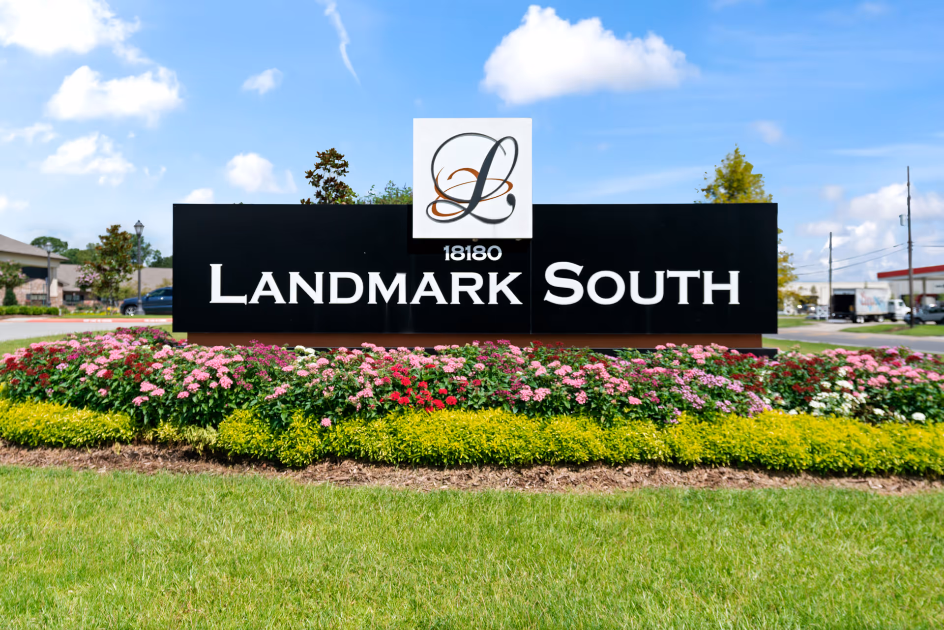 Outdoor view of a large black sign with white text reading '18180 Landmark South' surrounded by neatly trimmed green grass and colorful flower beds under a blue sky with some clouds.