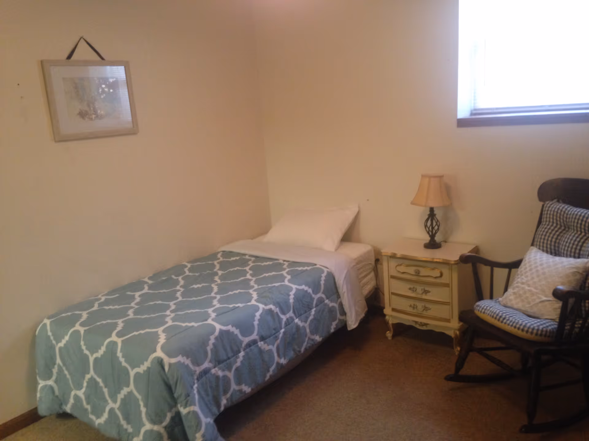 A small bedroom with a single bed covered with a blue and white patterned comforter and a white pillow. Next to the bed is a vintage-style nightstand with a small lamp on it. To the right of the nightstand is a cushioned wooden rocking chair with two pillows. A framed picture hangs on the wall above the bed, and there is a window with blinds letting in natural light.