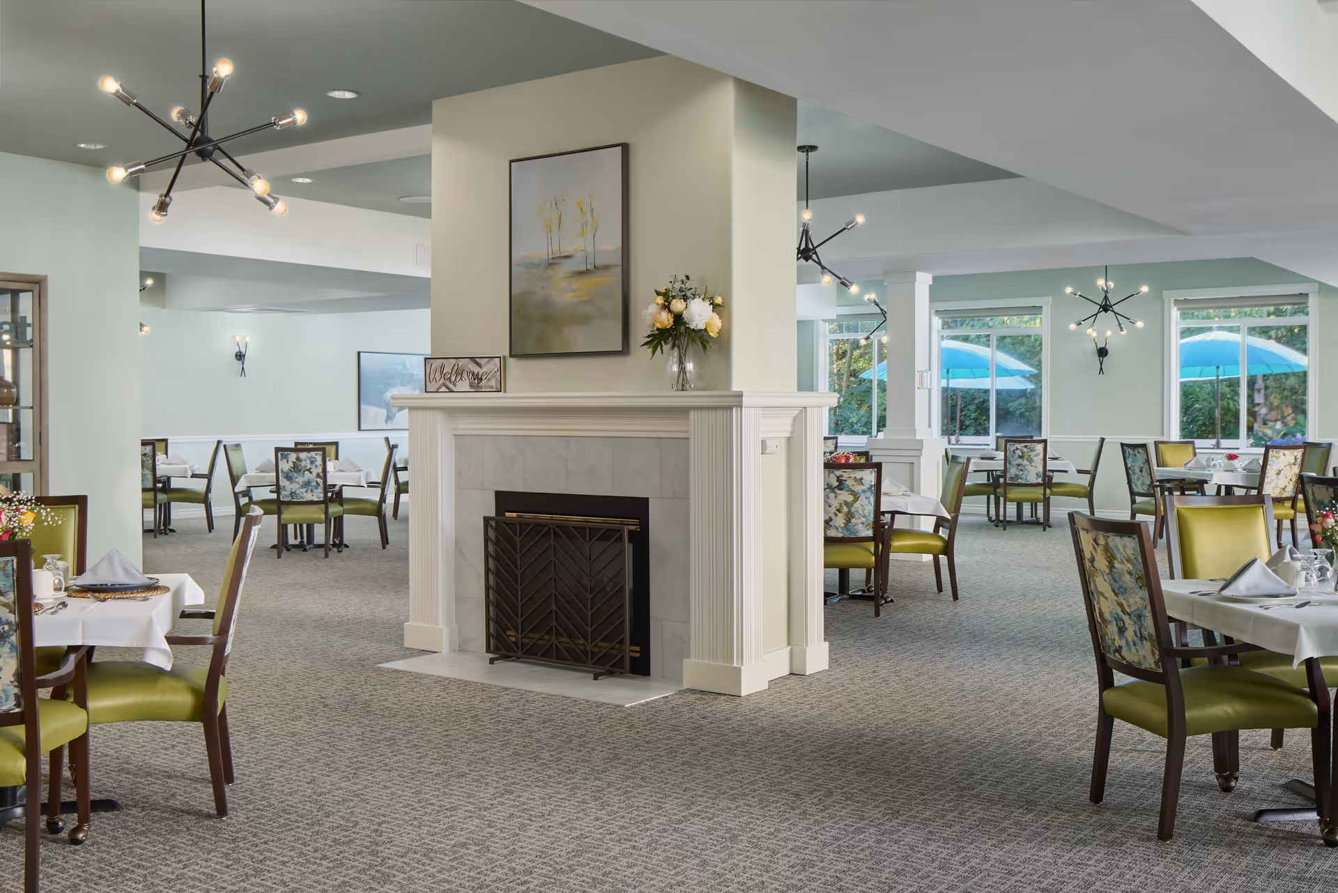 A spacious dining room in a senior living facility with multiple tables covered with white tablecloths, set with napkins and glassware. The room features green cushioned chairs with floral backs, a central fireplace with a painting and flowers on the mantel, modern light fixtures, and large windows showing blue umbrellas outside.