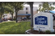 Exterior view of a senior living facility front with a sign, trees, and parked cars.