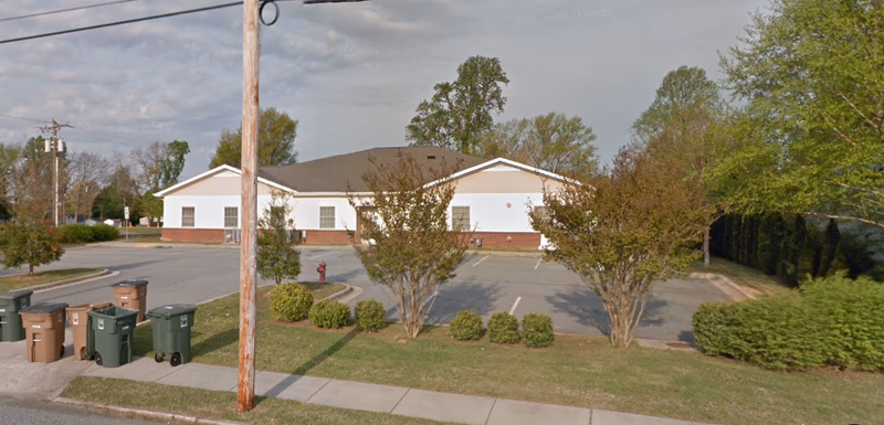Single-story white senior living facility with driveway, parking lot, and landscaped trees.