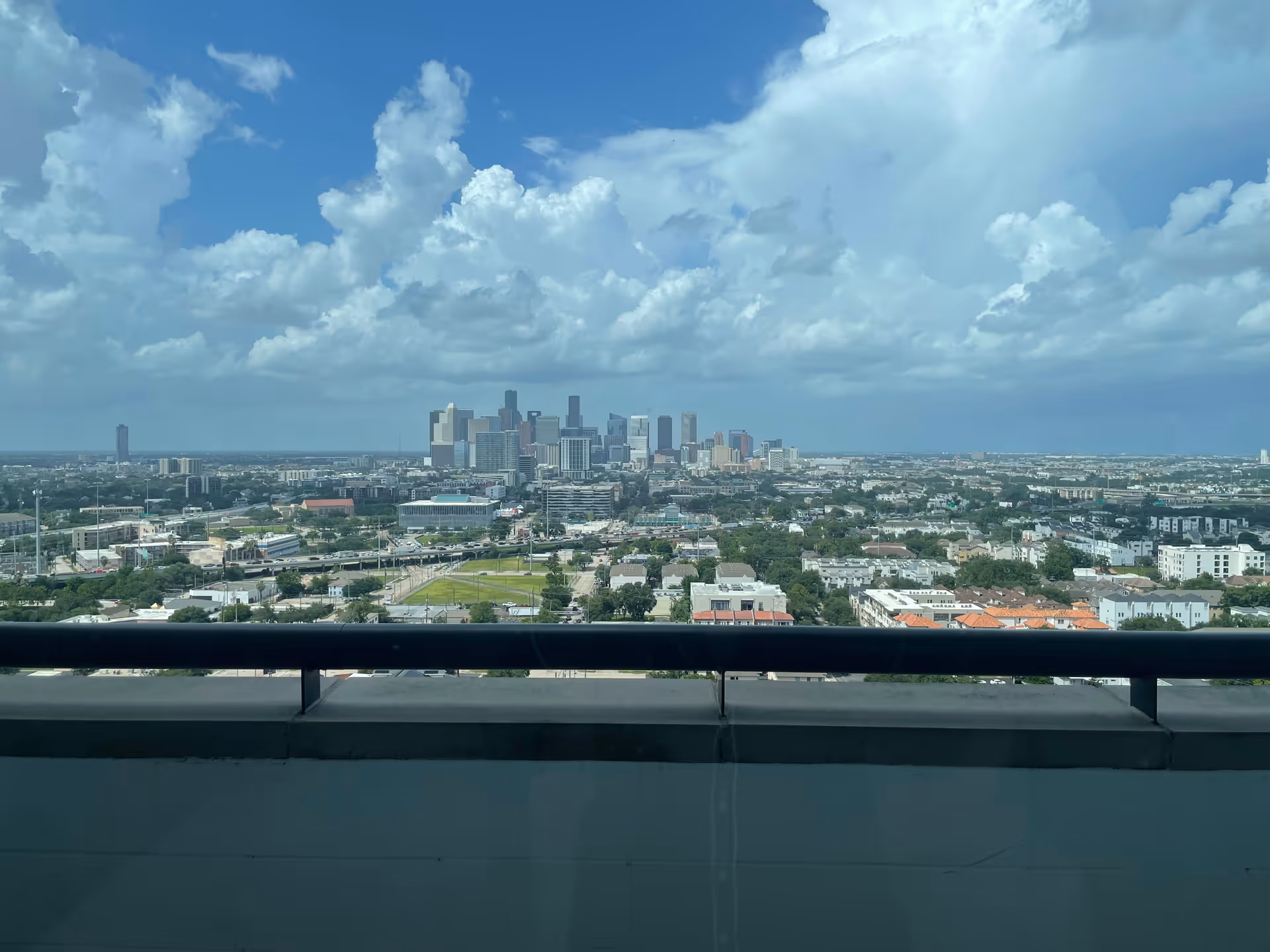 City skyline seen from a balcony railing with a cloudy blue sky above.