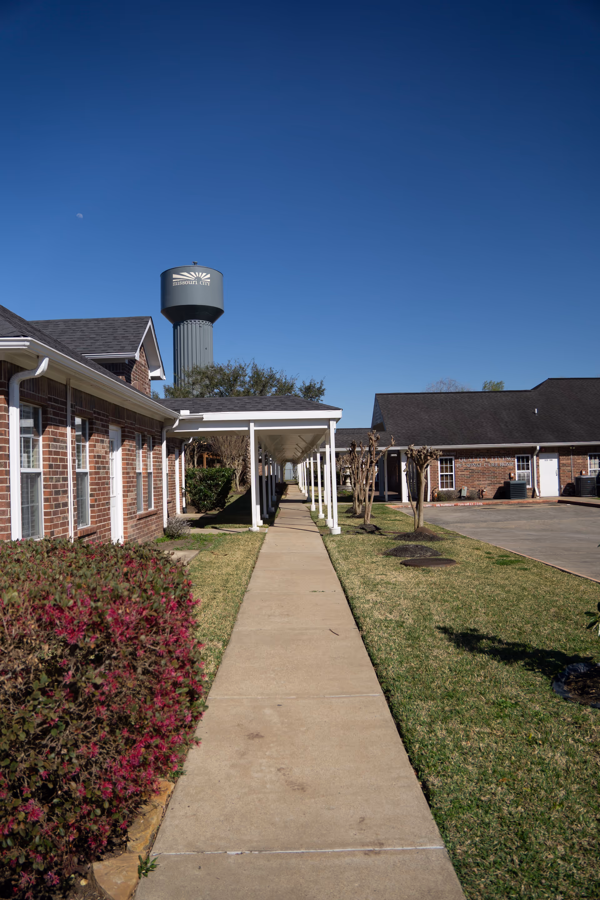 A concrete walkway with a covered colonnade between low brick buildings leading toward a distant water tower under a clear blue sky.