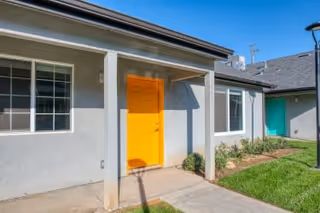 Exterior view of a single-story building with a bright yellow door, beige walls, and white-framed windows. There is a small covered porch supported by a column, a concrete walkway leading to the door, green grass, and a clear blue sky.