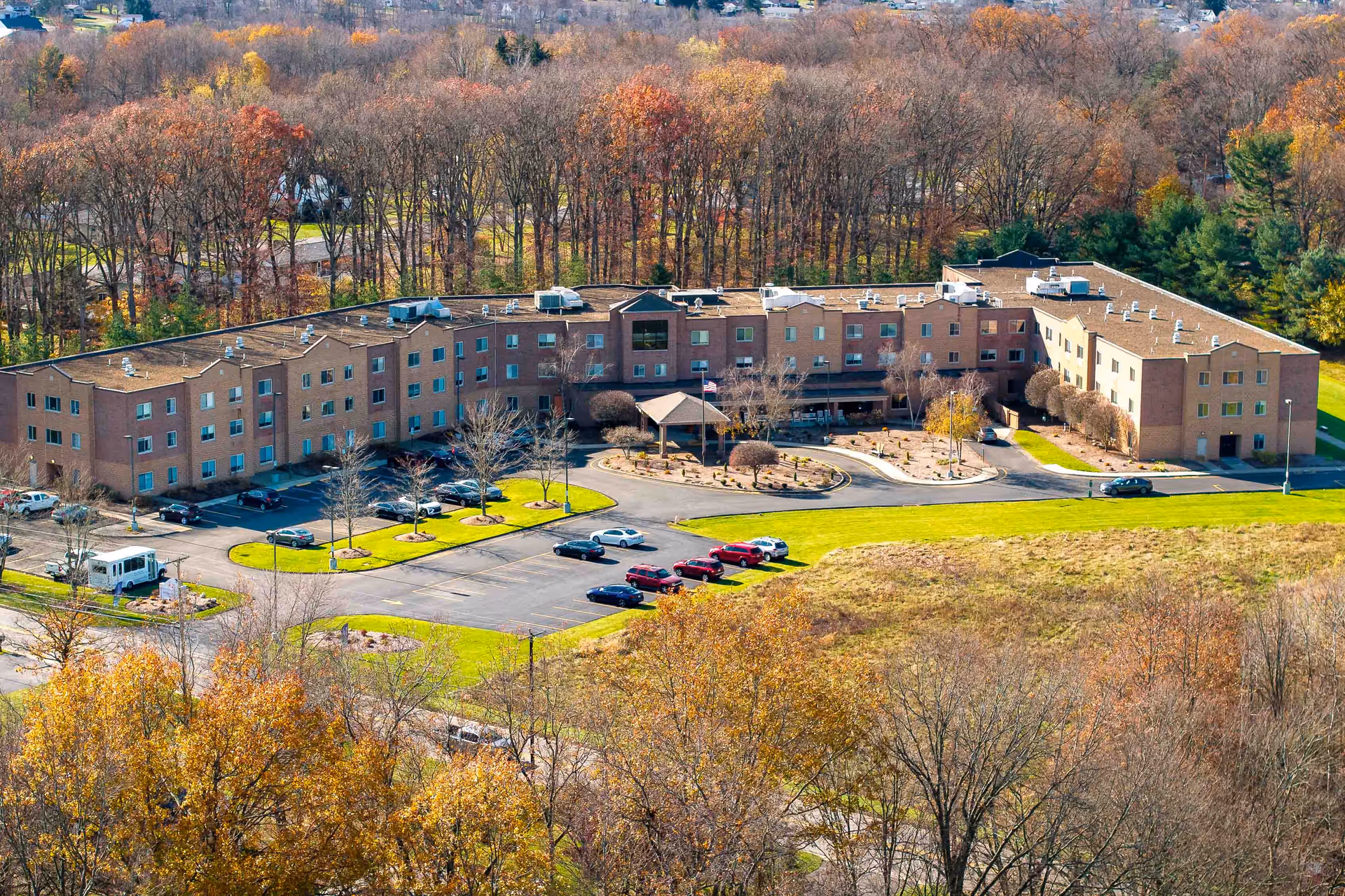 Aerial view of a large, three-story senior living facility named Whispering Oaks - A Provincial Senior Living Community, surrounded by trees with autumn foliage. The building has a brick exterior and a parking lot with several cars parked. The entrance features a covered drop-off area with a circular driveway.