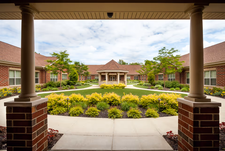 Covered walkway frames a brick senior living courtyard with yellow shrubs and a central pavilion.