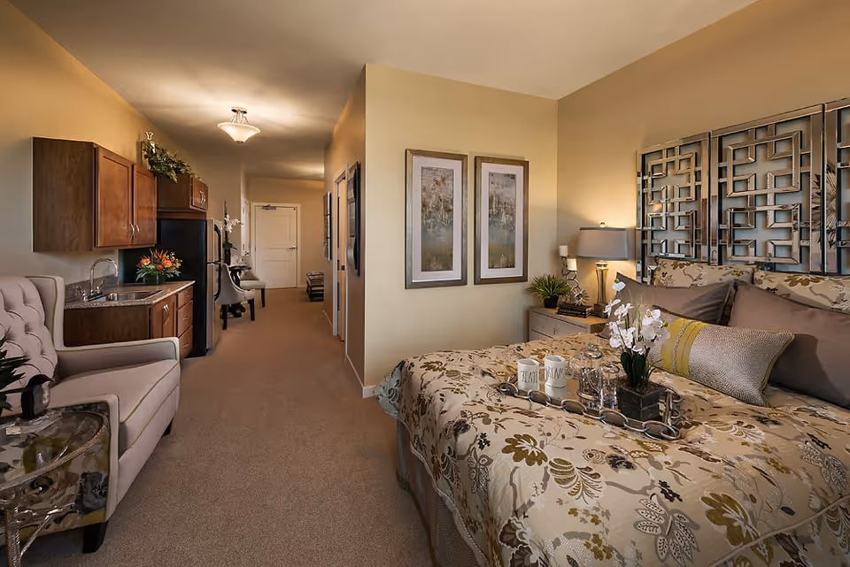 Cozy senior living studio featuring a decorated bed, bedside table and lamp, a small kitchenette and a seating area in a warm, neutral palette.