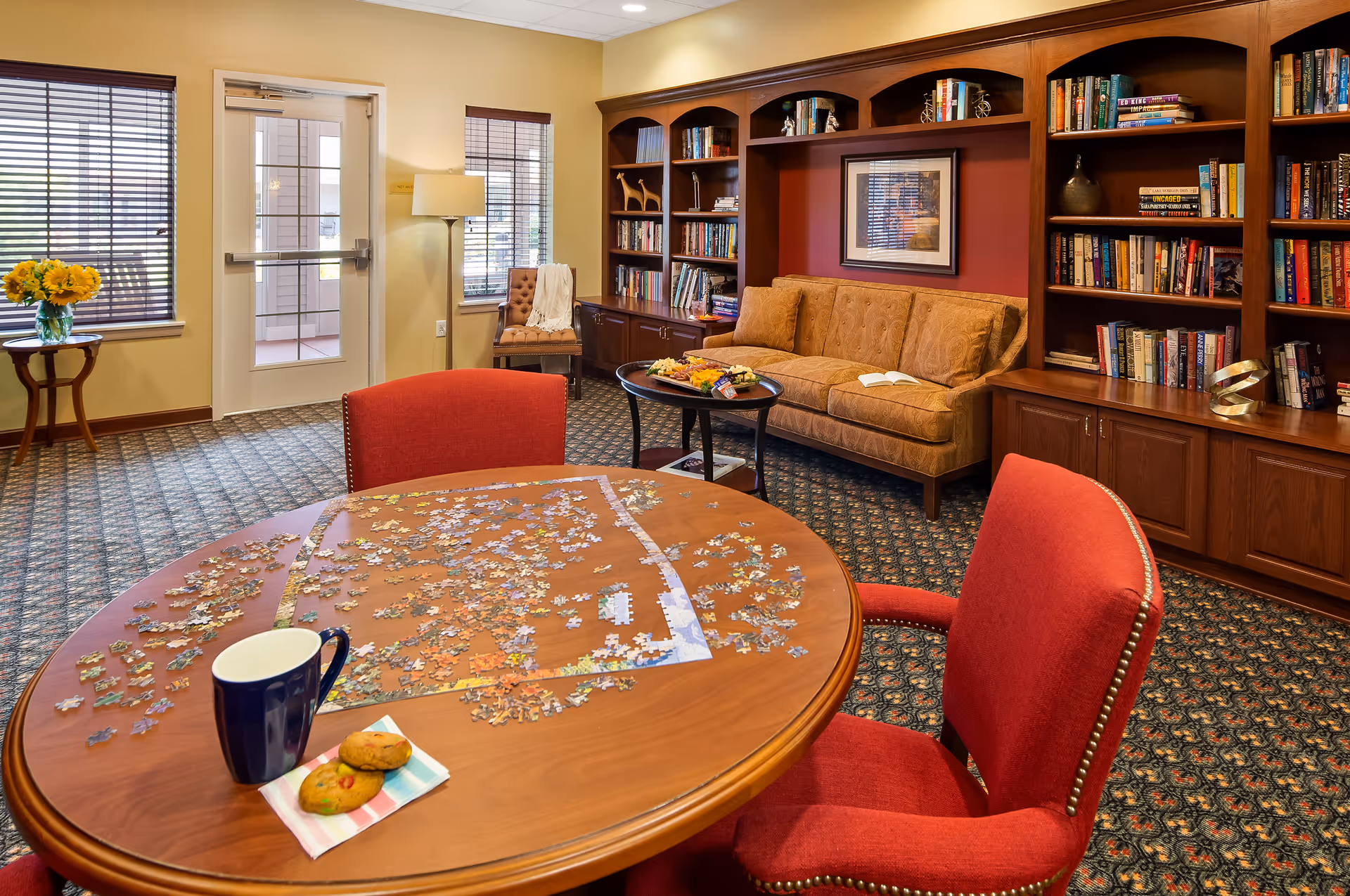 A cozy living room area with a round wooden table in the foreground covered with an incomplete jigsaw puzzle, a blue mug, and two cookies on a napkin. The room features red upholstered chairs around the table, a beige patterned sofa against a wall with built-in wooden bookshelves filled with books and decorative items, a small round table with a platter of snacks, a framed picture on the wall, and two windows with blinds letting in natural light. A door with glass panels and a small side table with a vase of sunflowers are also visible.
