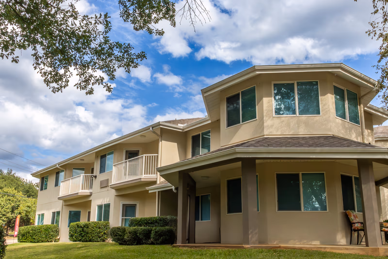 Exterior view of a two-story beige building with multiple windows and small balconies, surrounded by green bushes and grass under a partly cloudy blue sky.