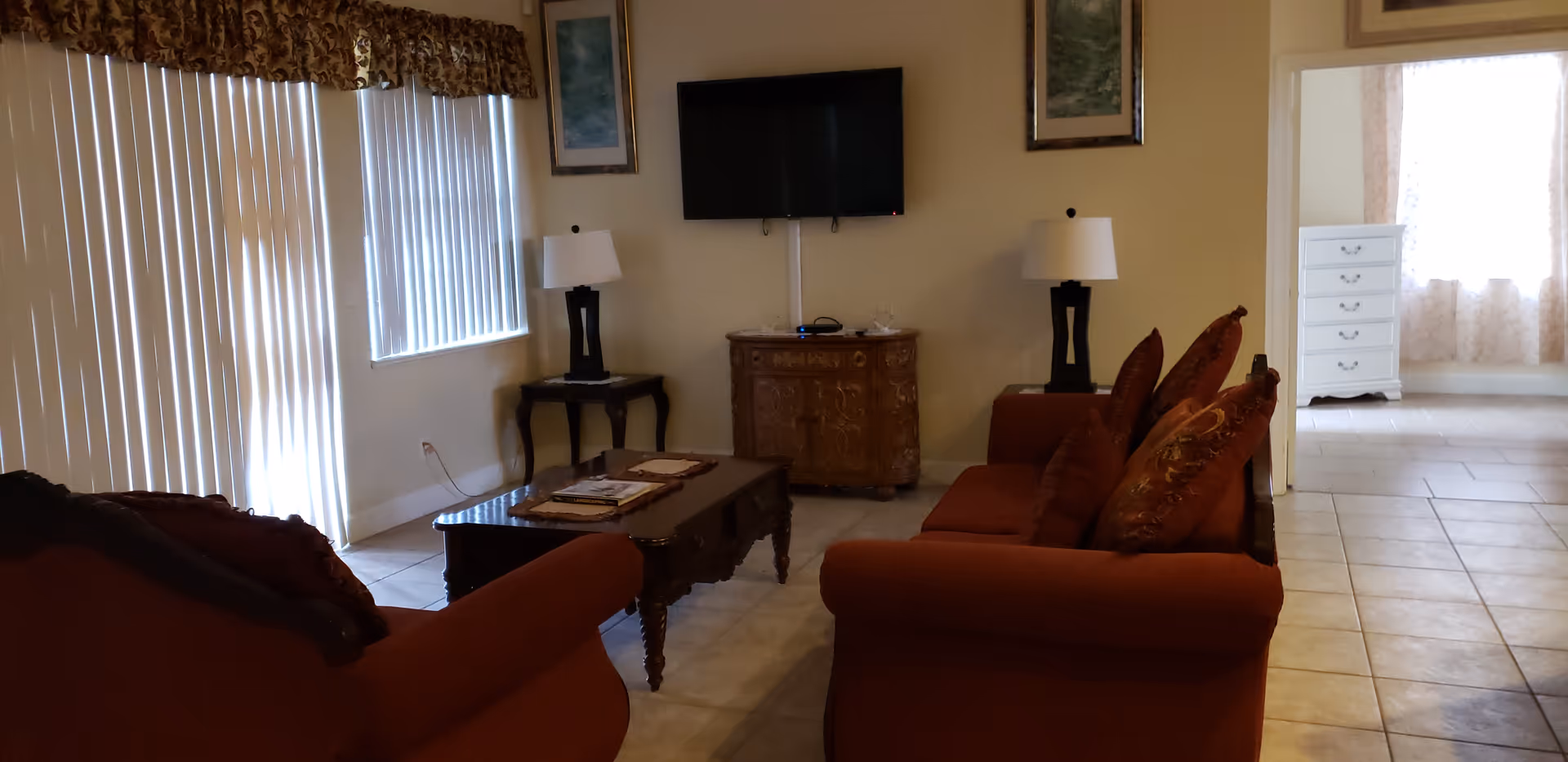 Cozy living room with red sofas, a coffee table, wall-mounted TV, side lamps, and vertical blinds over a sliding door.