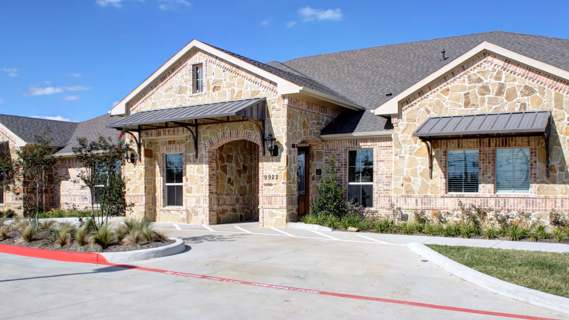 Exterior view of a stone and brick building with a covered entrance, windows with awnings, and landscaped greenery in front under a clear blue sky.