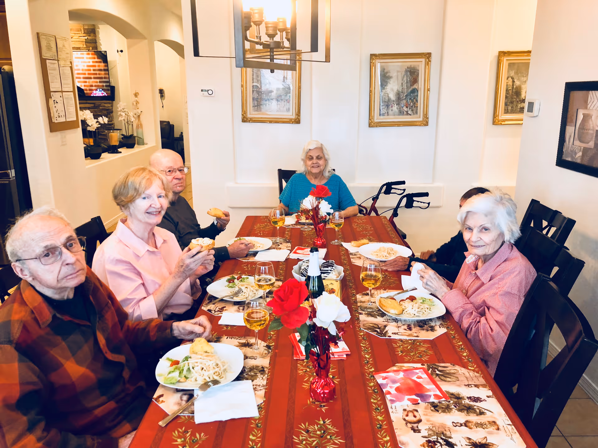A group of six elderly people sitting around a dining table enjoying a meal together in a well-lit room. The table is covered with a red tablecloth and decorated with red and white flowers. Plates of food, glasses of a yellow beverage, and bread rolls are visible on the table. The room has framed artwork on the walls and a chandelier overhead.