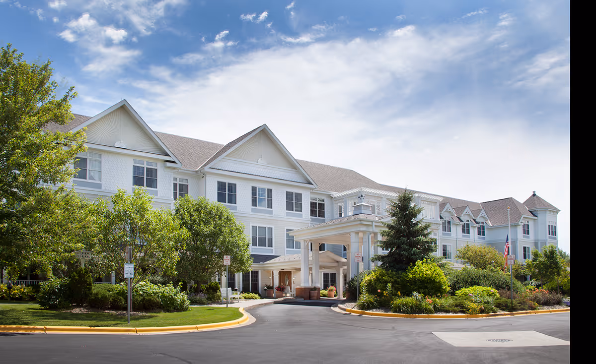 White multi-story senior living building with a porte-cochere entrance and lush landscaping.