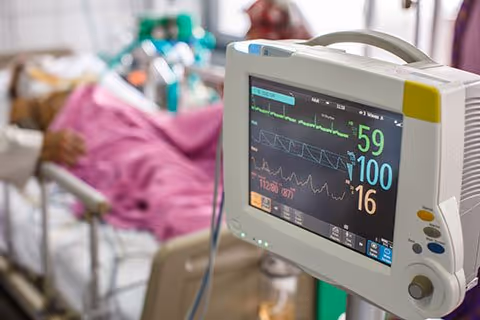 A close-up view of a medical monitor displaying vital signs including heart rate, oxygen saturation, and respiratory rate, with a patient lying in a hospital bed covered with a pink blanket in the background.