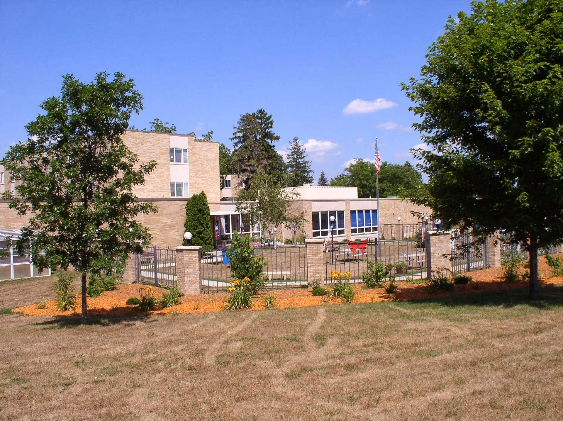 Exterior view of Trempealeau County Health Care Center showing a brick building with multiple windows, surrounded by trees, a fenced garden area with flowers, and an American flag on a flagpole under a clear blue sky.