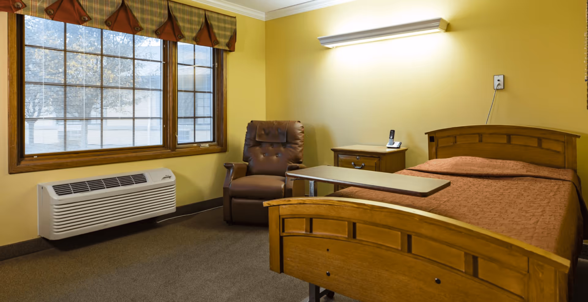 A simple room with a single wooden bed covered with a brown quilt, a brown leather recliner chair, a wooden nightstand with a cordless phone, a large window with blinds and a valance, and a wall-mounted air conditioning unit below the window.