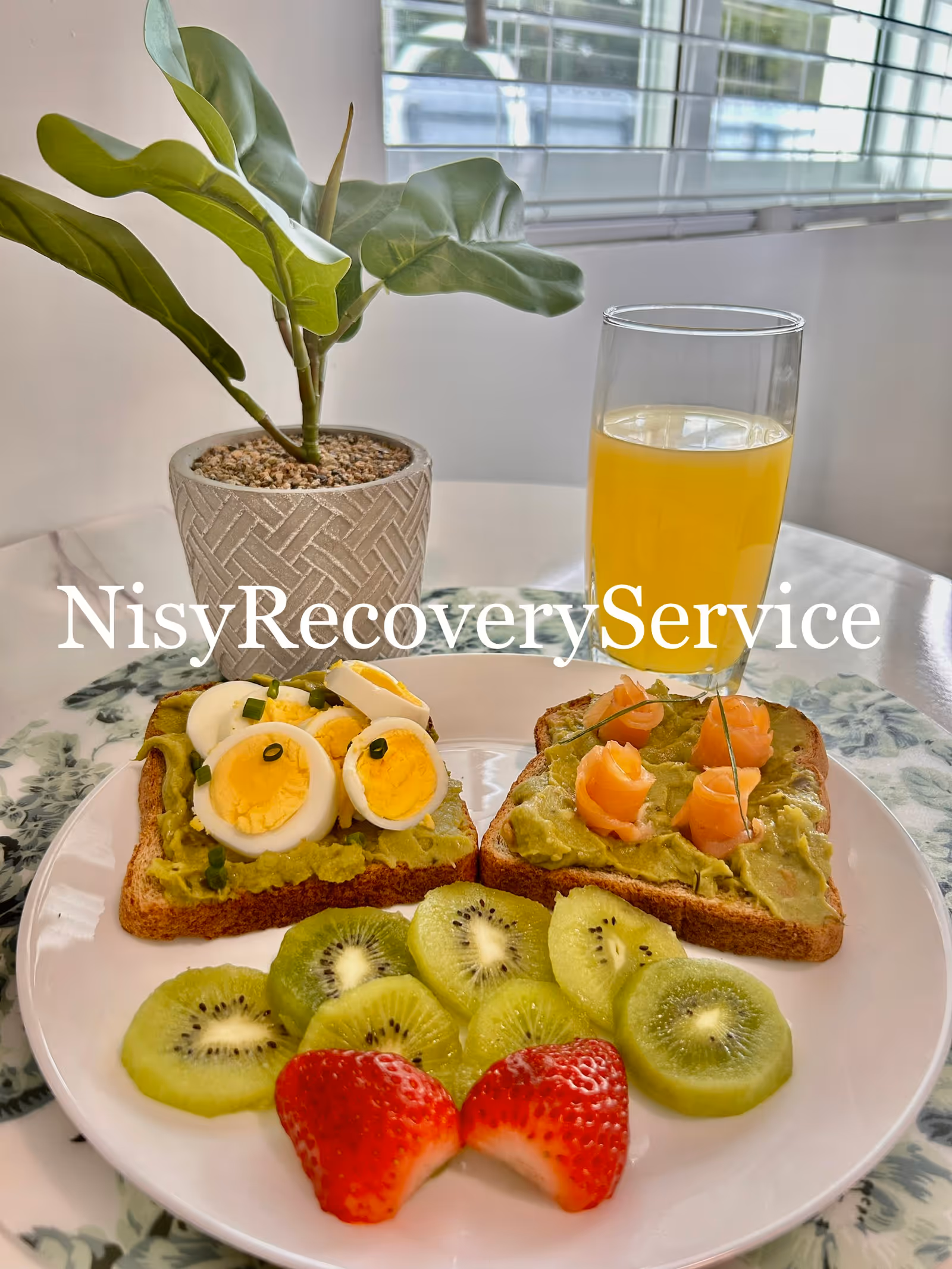 Indoor dining table scene with a plant, juice, avocado toasts, and kiwi slices.