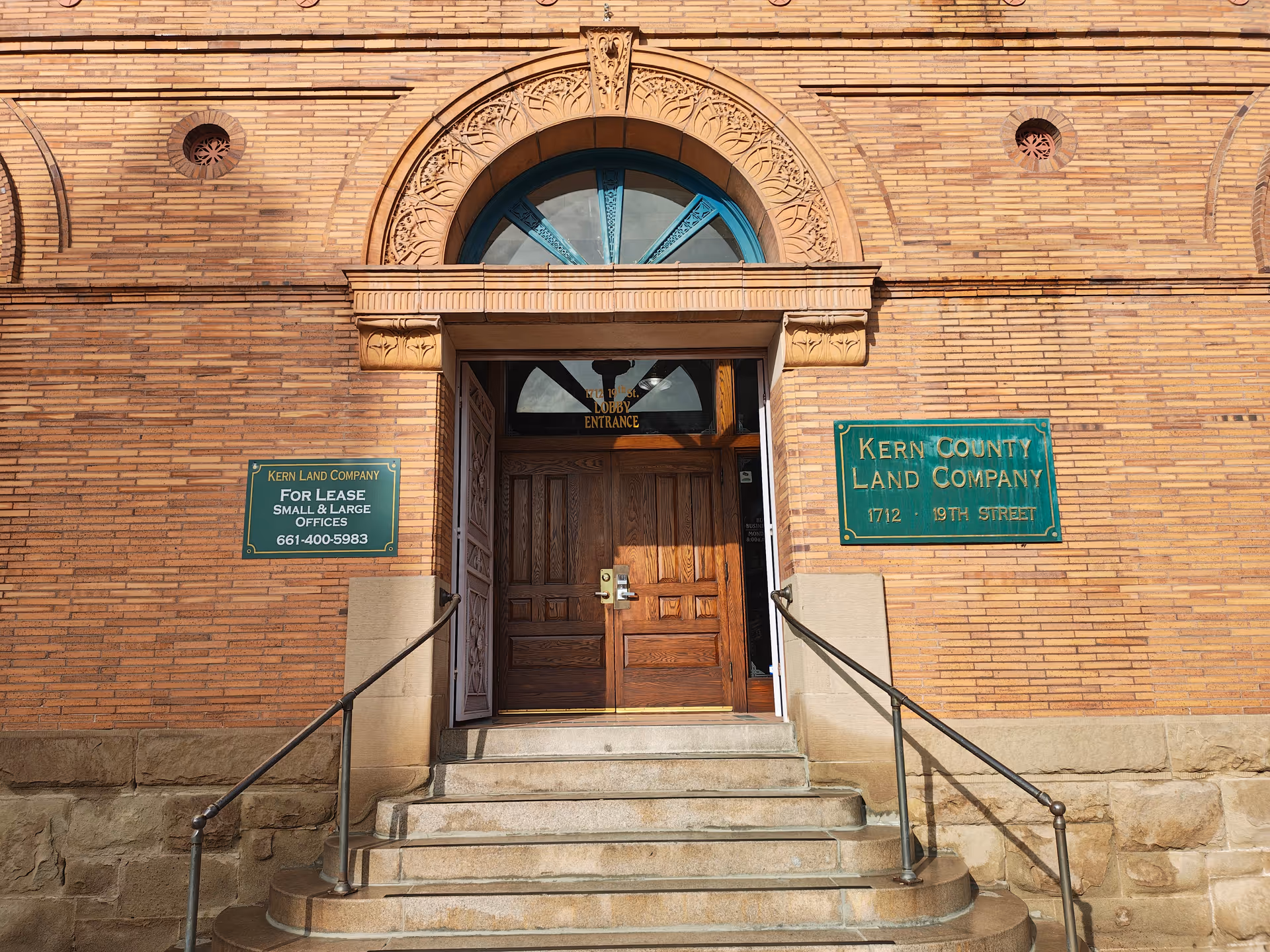 Brick building facade with arched window, wooden doors, steps, and green plaques at entrance.