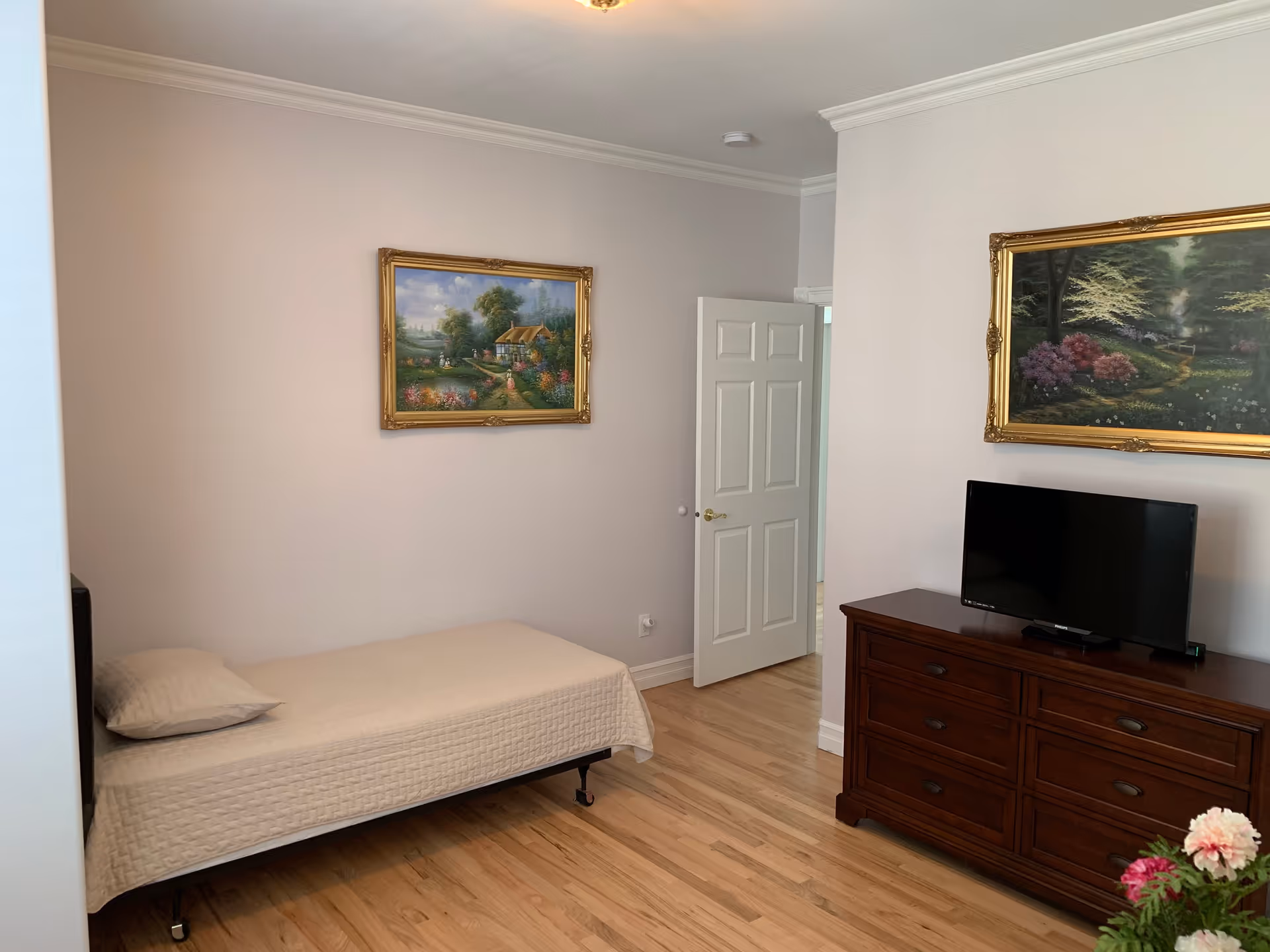 A simple bedroom with a single bed covered in a beige quilt and a matching pillow. The room has light-colored walls with two framed landscape paintings hanging on them. There is a wooden dresser with a flat-screen TV on top. The floor is wooden, and a white door is partially open, revealing a glimpse of another room.