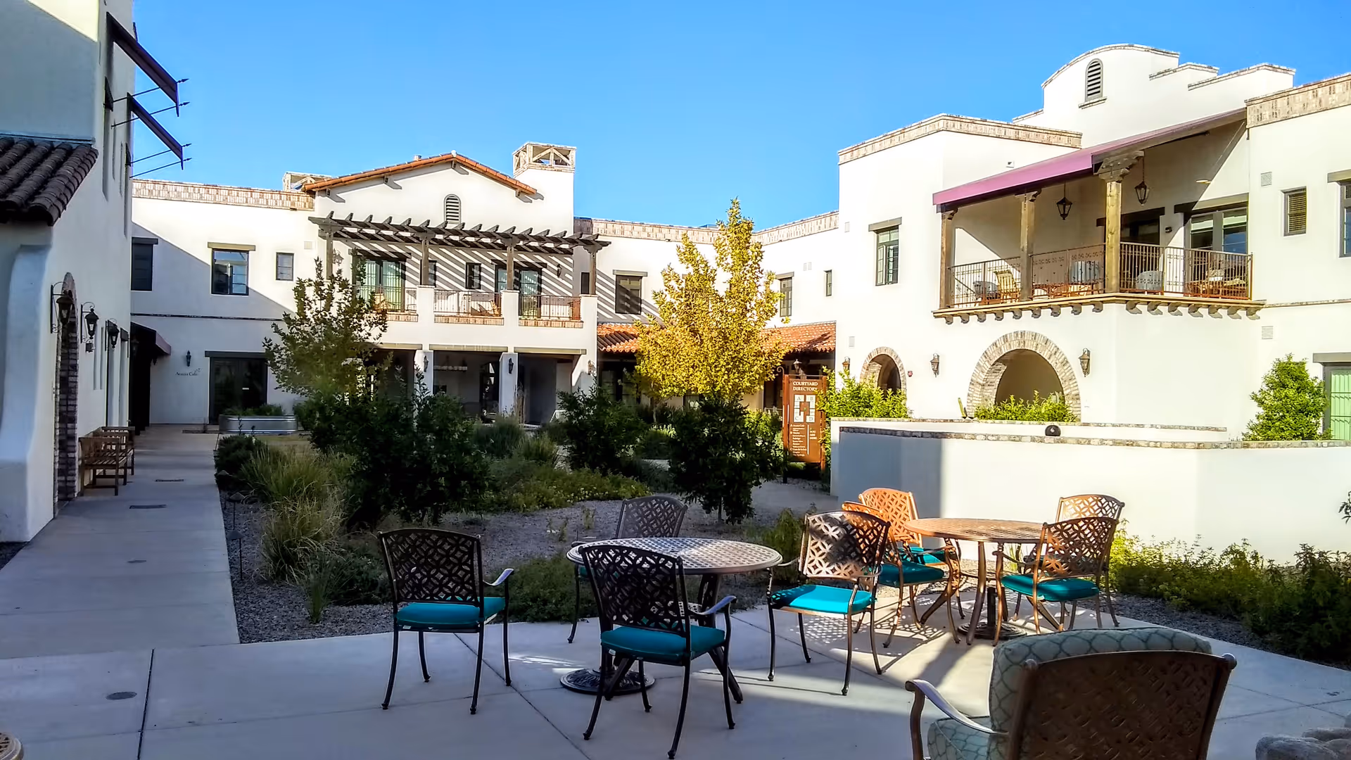 An outdoor courtyard with white stucco buildings, stone arches, and metal tables with teal chairs.