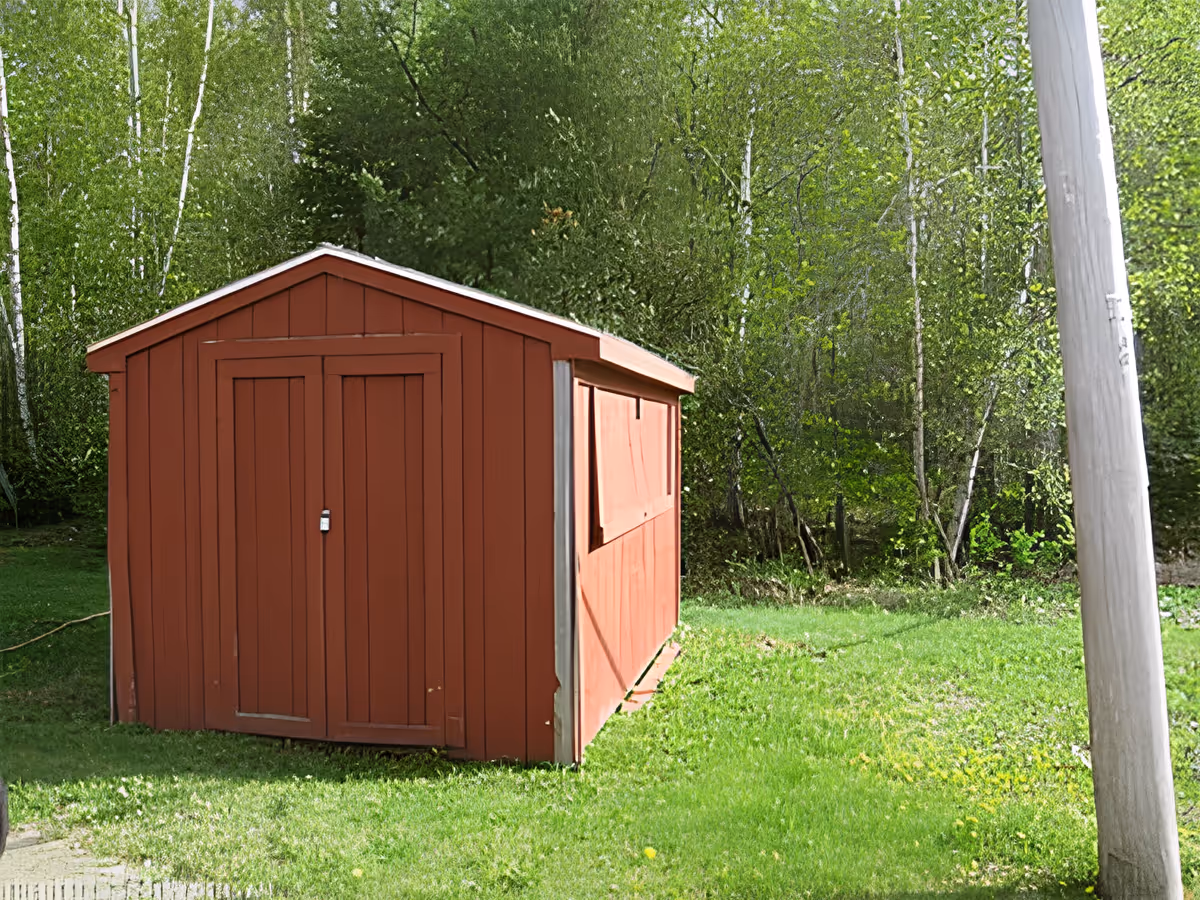 A small red wooden shed with double doors and a padlock, situated on a grassy area with dense green trees and bushes in the background. A wooden utility pole is visible on the right side of the image.