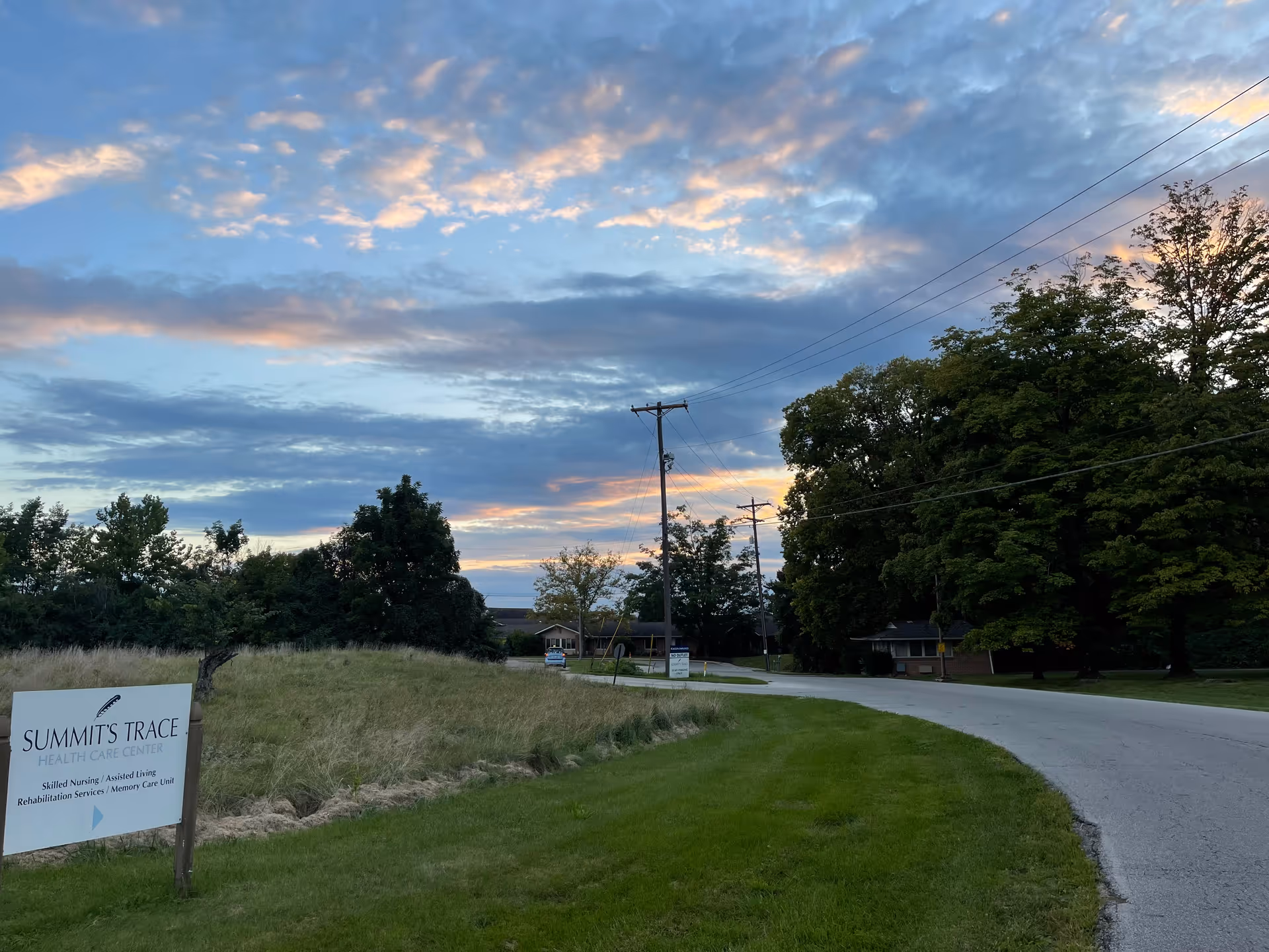Sunset sky over a curved road with Summit's Trace sign beside green lawn.