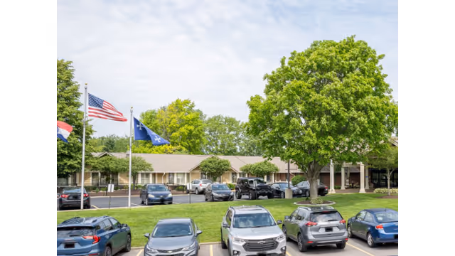 Exterior view of a senior care facility with parked cars and flags along the front.