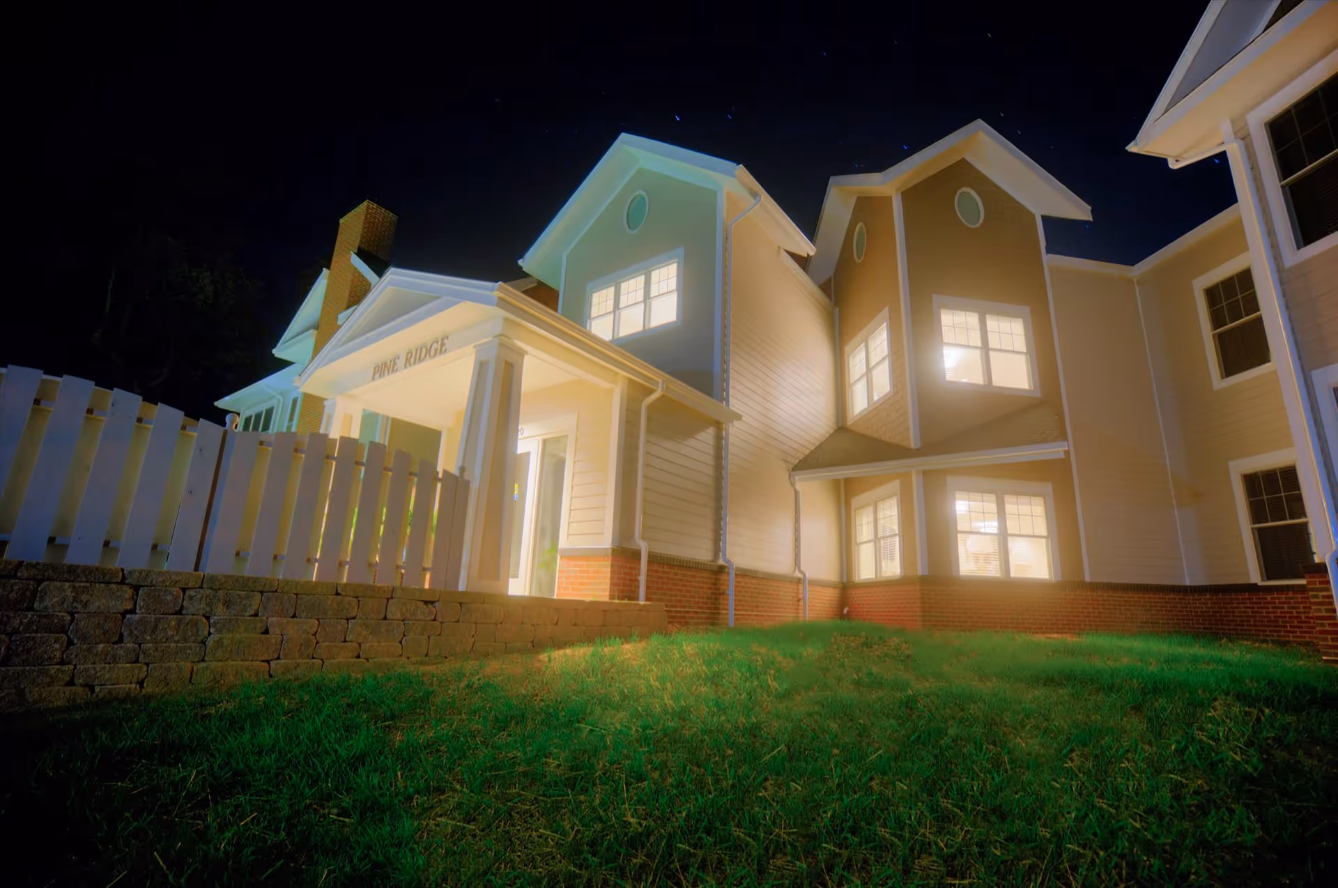 Night view of a well-lit residential building with multiple windows, a white picket fence, and a small grassy area in front. The building has a sign that reads 'PINE RIDGE' above the entrance.