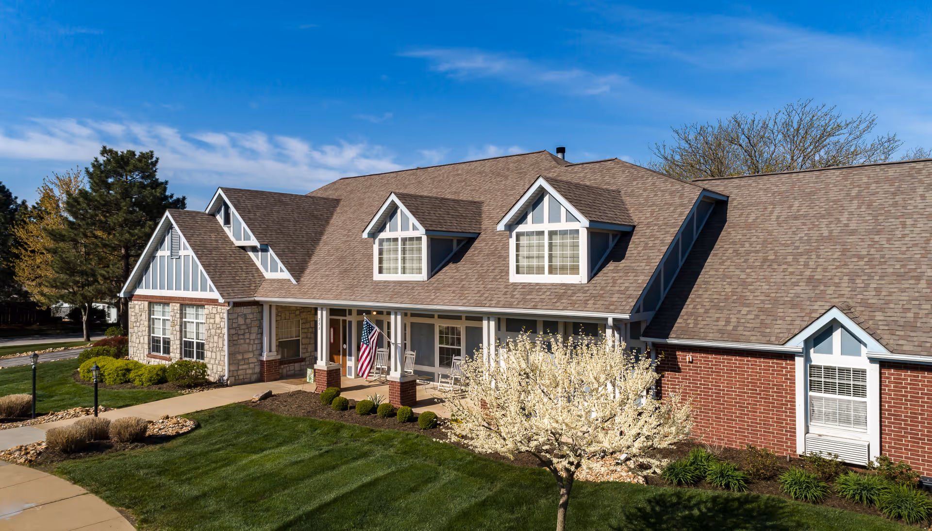 Exterior view of a single-story assisted living facility with a combination of brick and stone facade, multiple dormer windows, a covered porch with rocking chairs, an American flag, well-maintained landscaping, and a clear blue sky.
