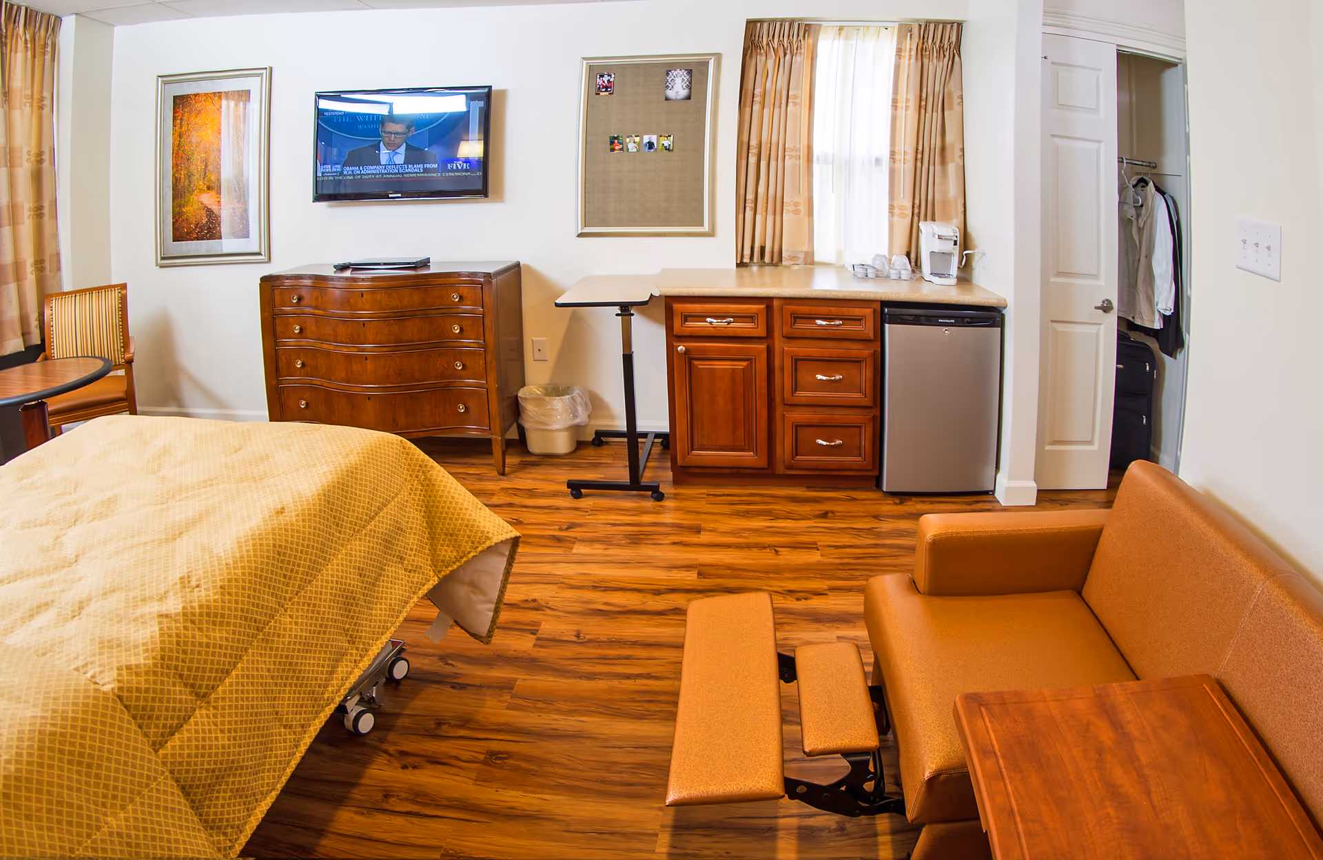 Interior senior living suite with a bed, couch, kitchenette, dresser, TV, and window curtains.