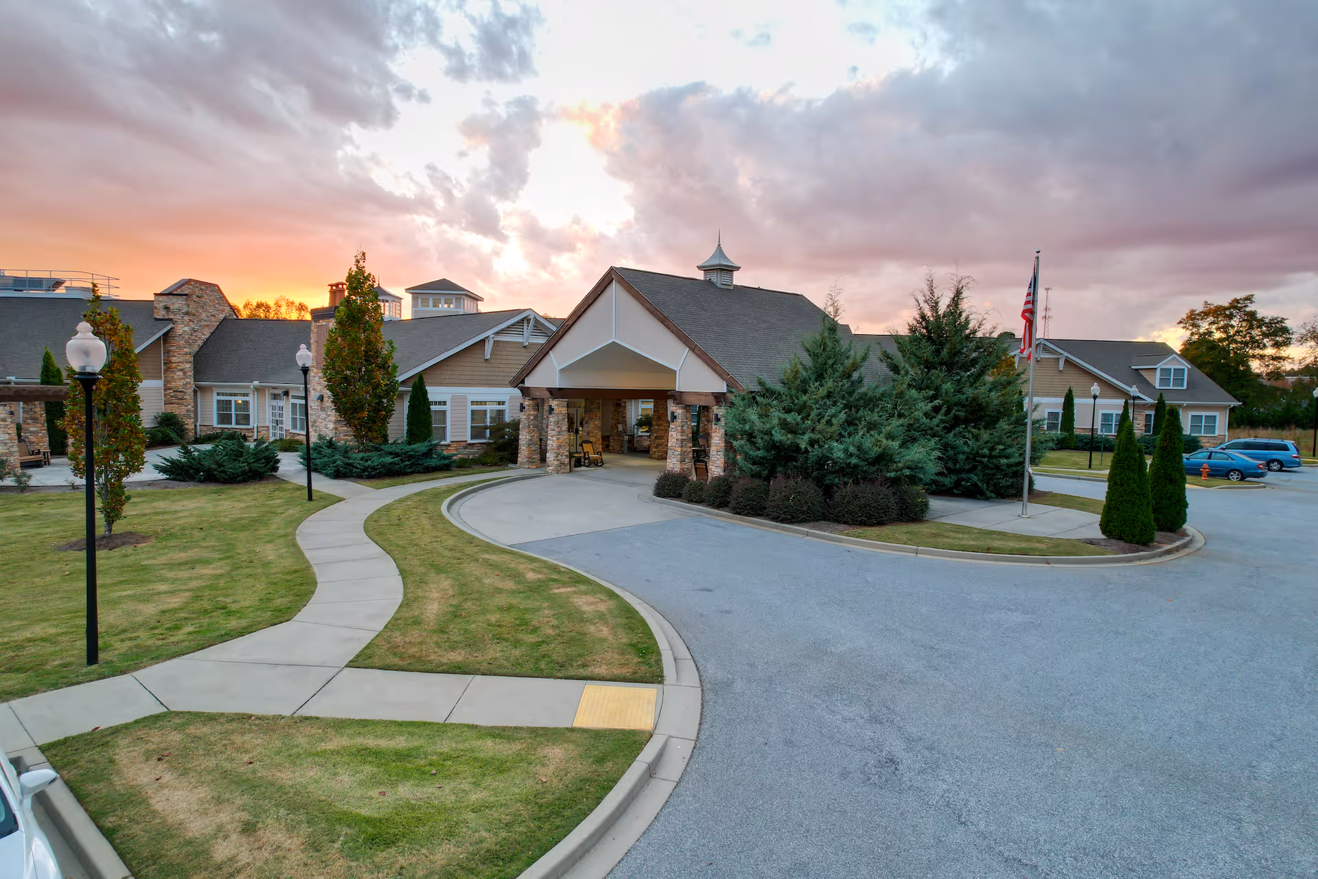 Exterior view of the Spring Park senior living facility at sunset, showing a driveway leading to a covered entrance with stone pillars, surrounded by well-maintained landscaping including trees, shrubs, and a lawn, with a few parked cars and an American flag on a flagpole.