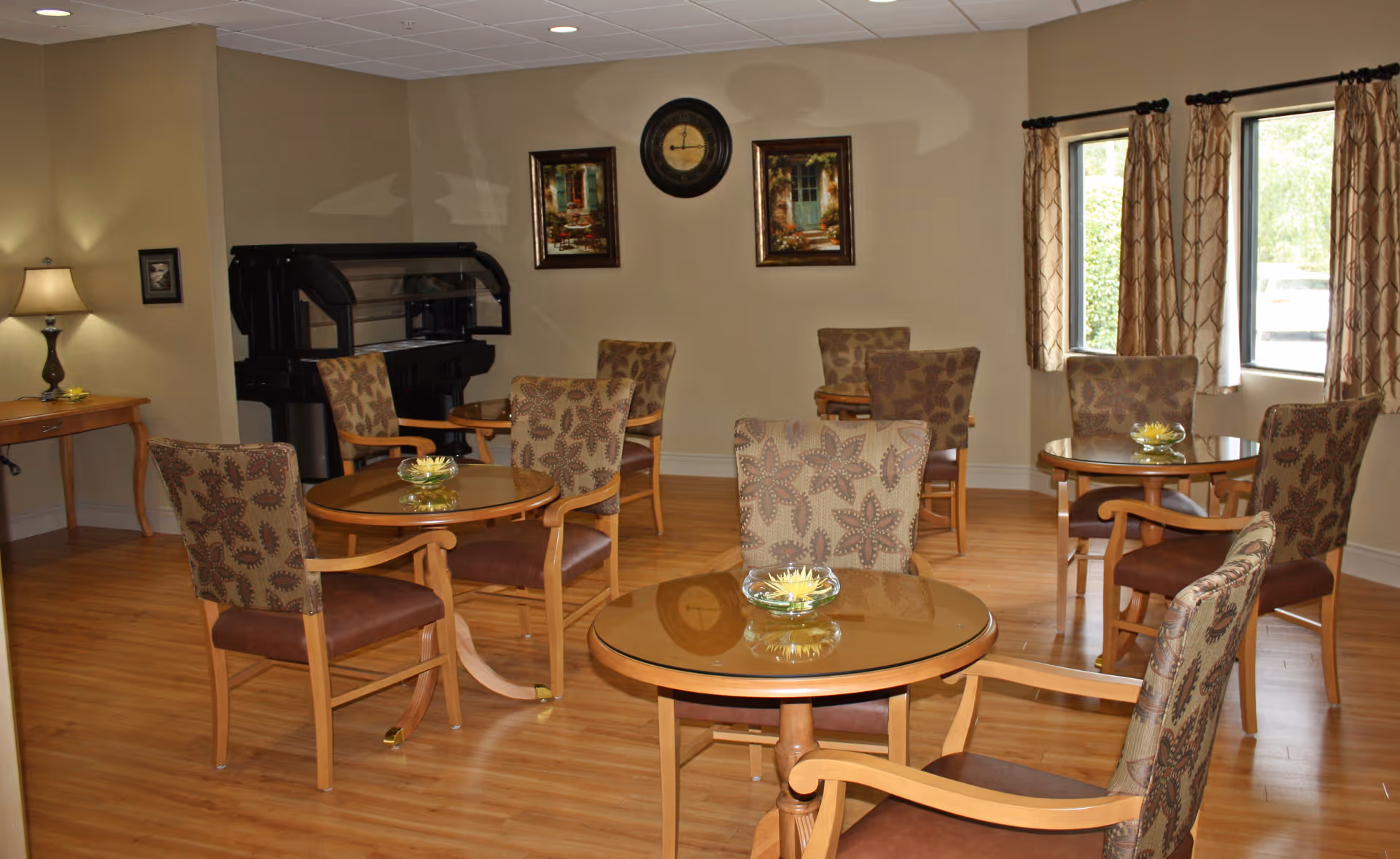 Well-lit common room with round glass-topped tables and patterned chairs on hardwood floors, a piano against the wall, framed art, and windows with curtains.