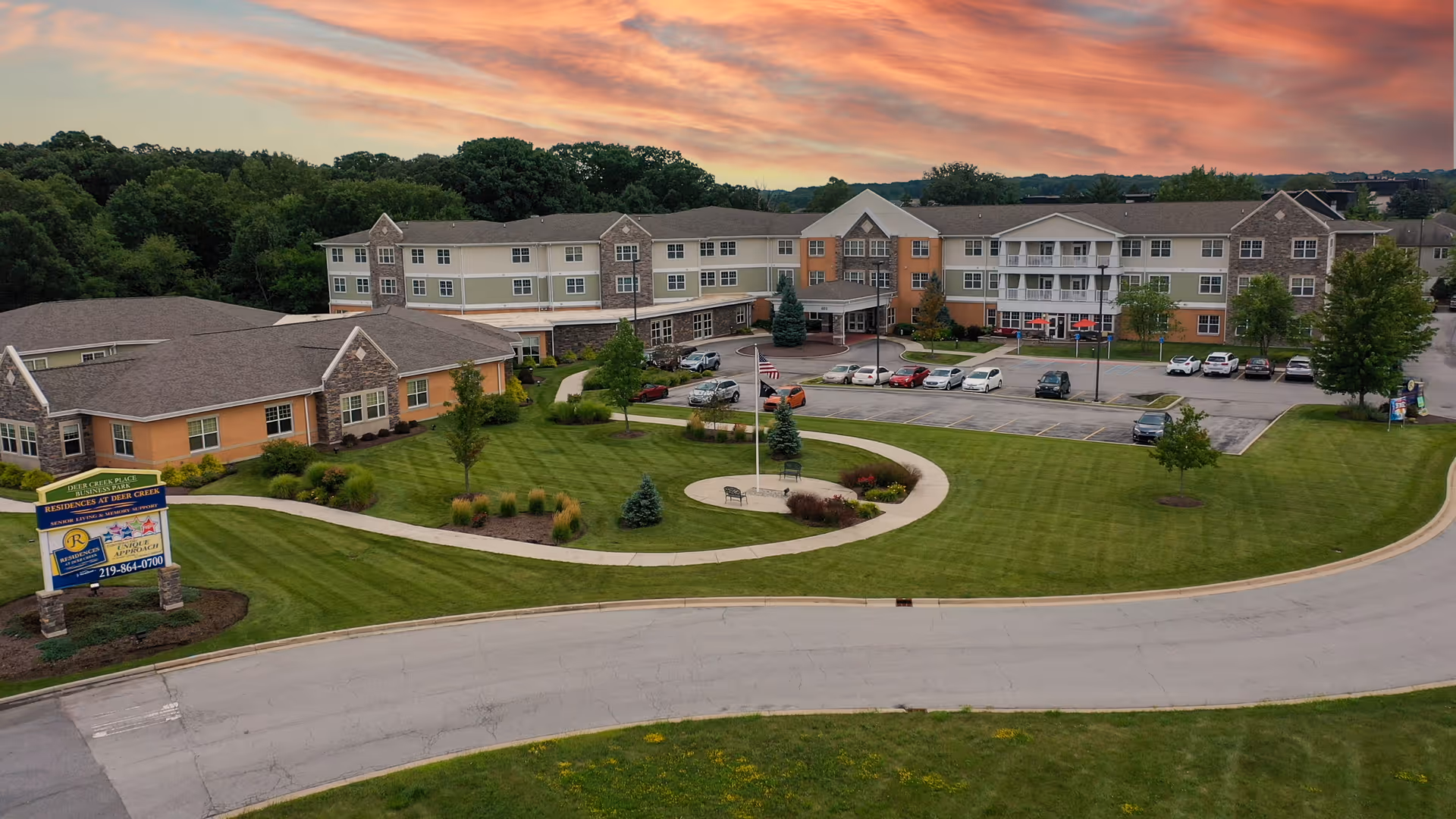 Aerial view of Residences At Deer Creek senior living facility during sunset, showing a large multi-story building with a parking lot, well-maintained green lawns, trees, and a circular walkway with benches and a flagpole in the center.