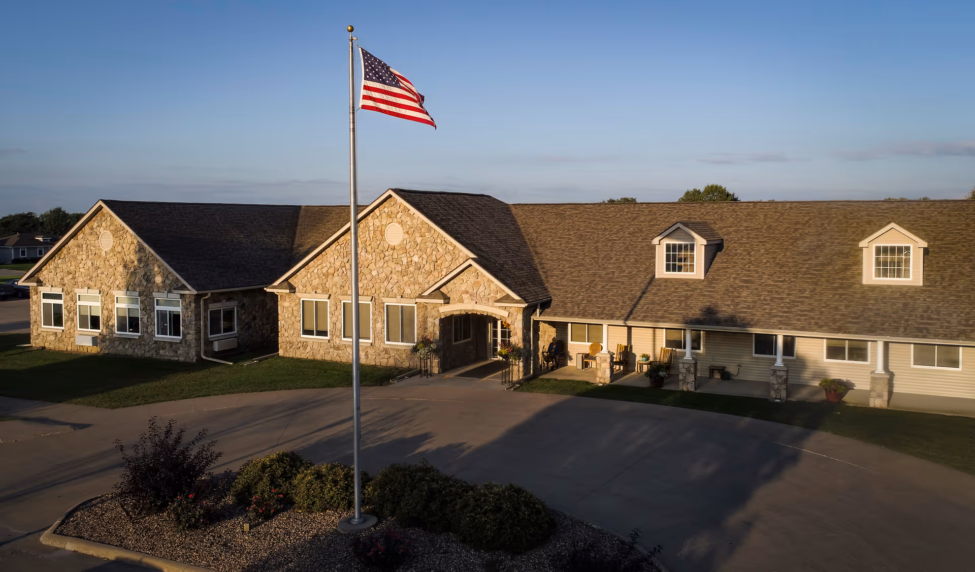 Exterior view of a single-story assisted living facility building with stone and siding facade, a central entrance, and an American flag on a flagpole in front. The building is surrounded by a paved driveway and landscaped greenery under a clear blue sky.