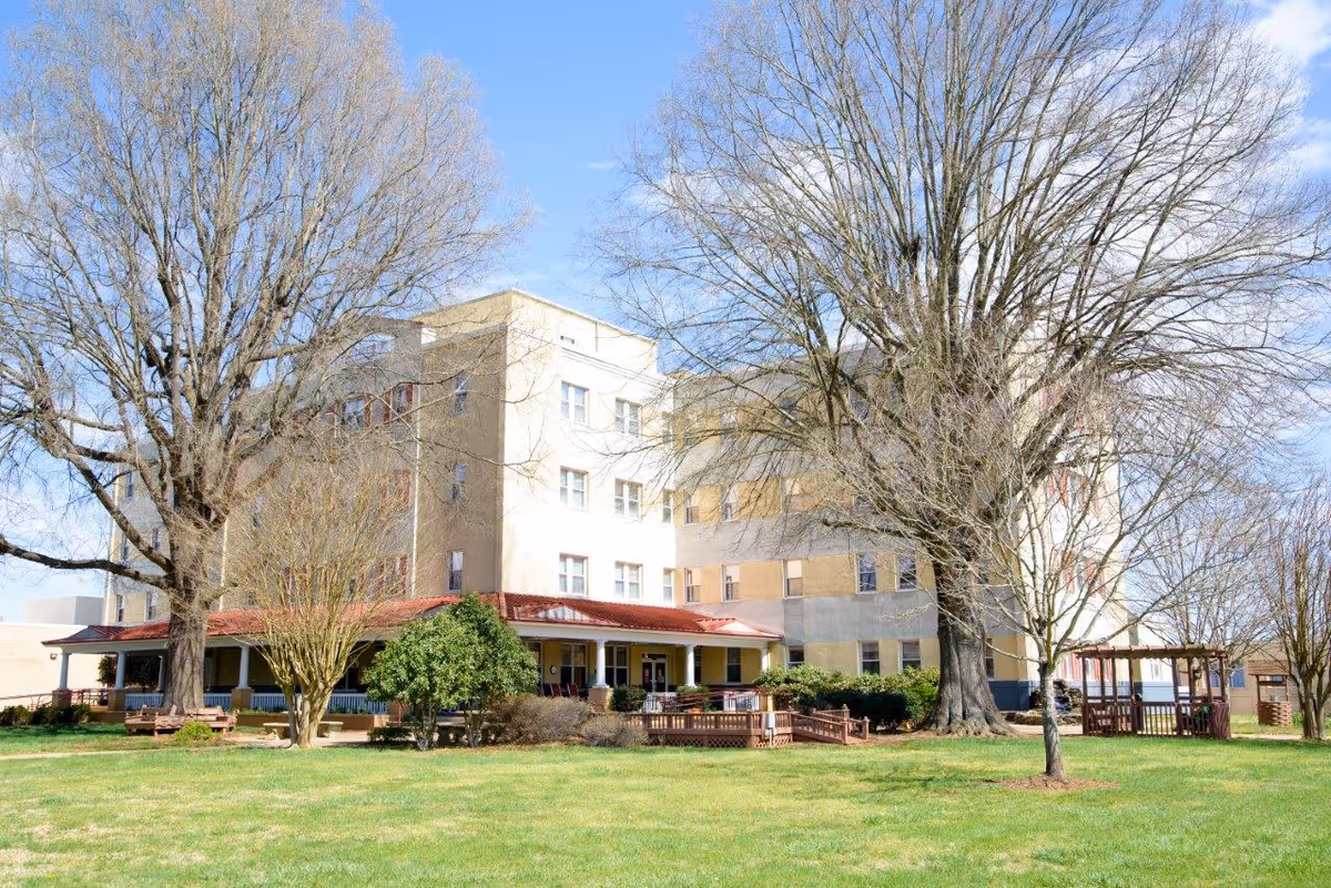 Exterior view of a multi-story senior living building with a covered porch, gazebo, benches, and large leafless trees on a grassy lawn.