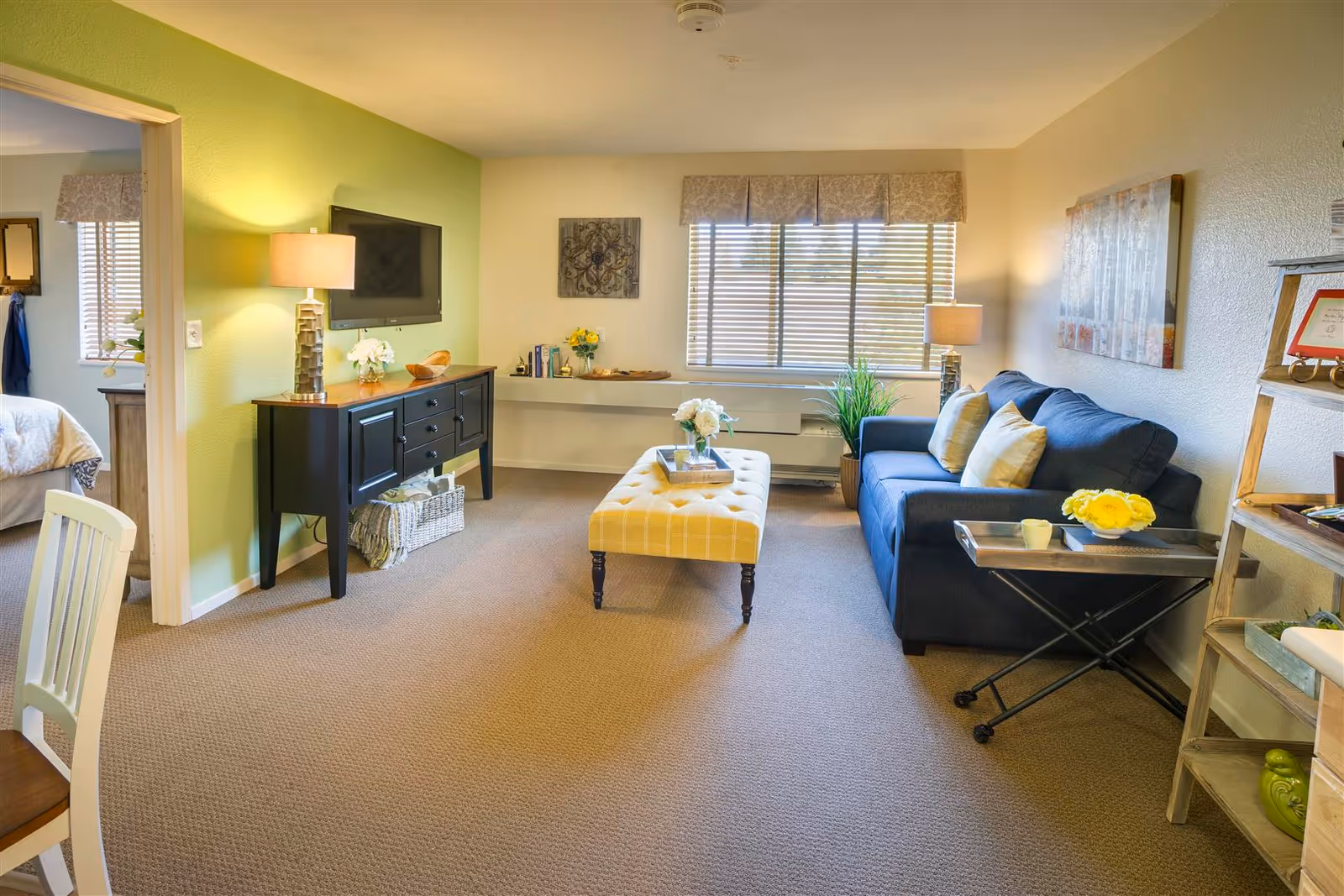 Bright living room in a senior living facility with couch, coffee table, TV, and windows.