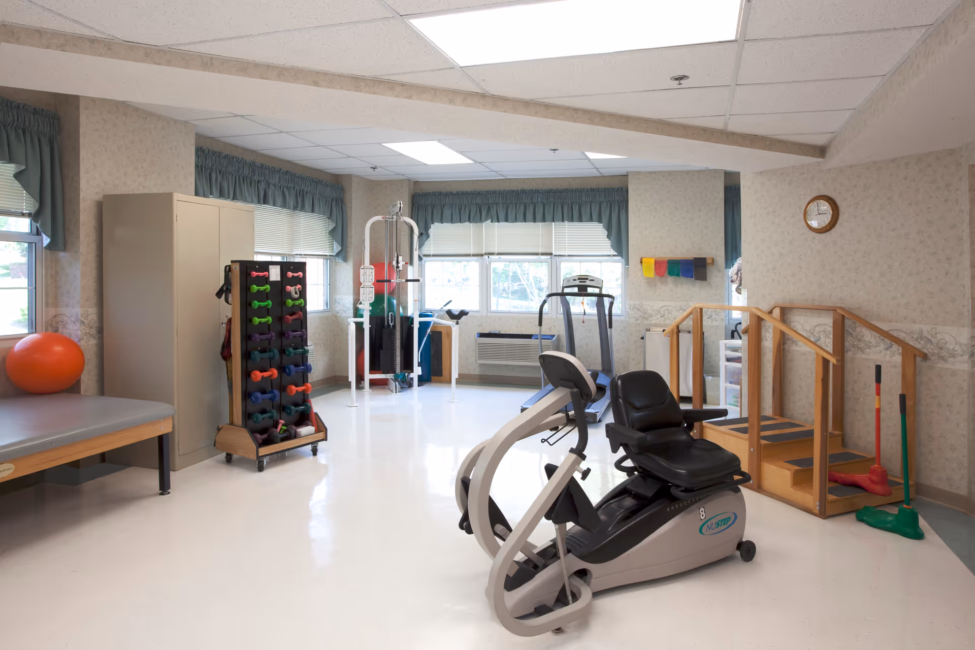 A well-lit physical therapy or exercise room with various equipment including a recumbent exercise bike, a treadmill, a rack of colorful dumbbells, a therapy table with an orange exercise ball, and a set of wooden stairs with handrails for rehabilitation exercises. The room has large windows with green valances and light-colored walls with a clock mounted on one wall.