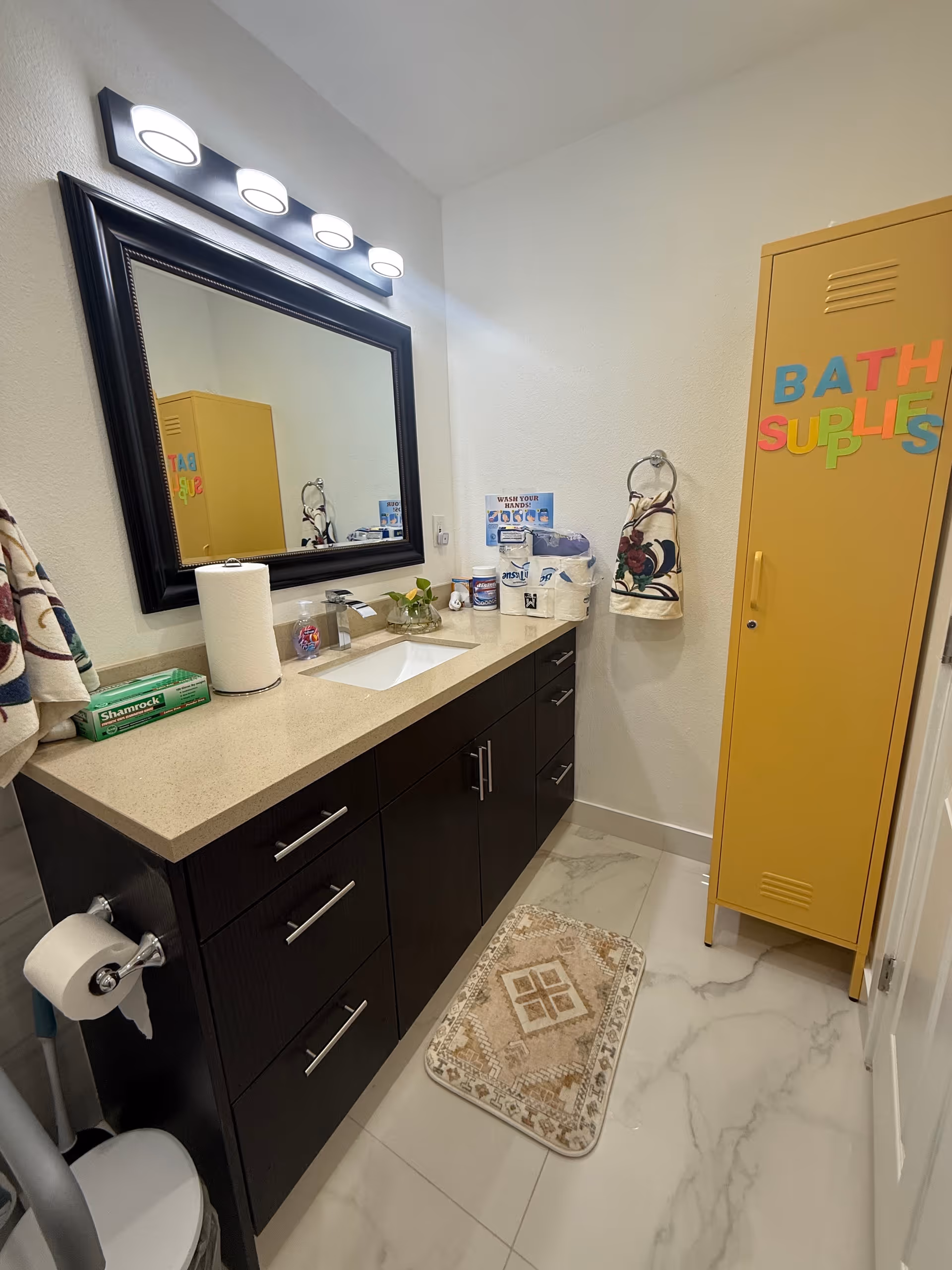 Sleek bathroom with dark vanity, large mirror, yellow storage cabinet, and marble floor tiles.
