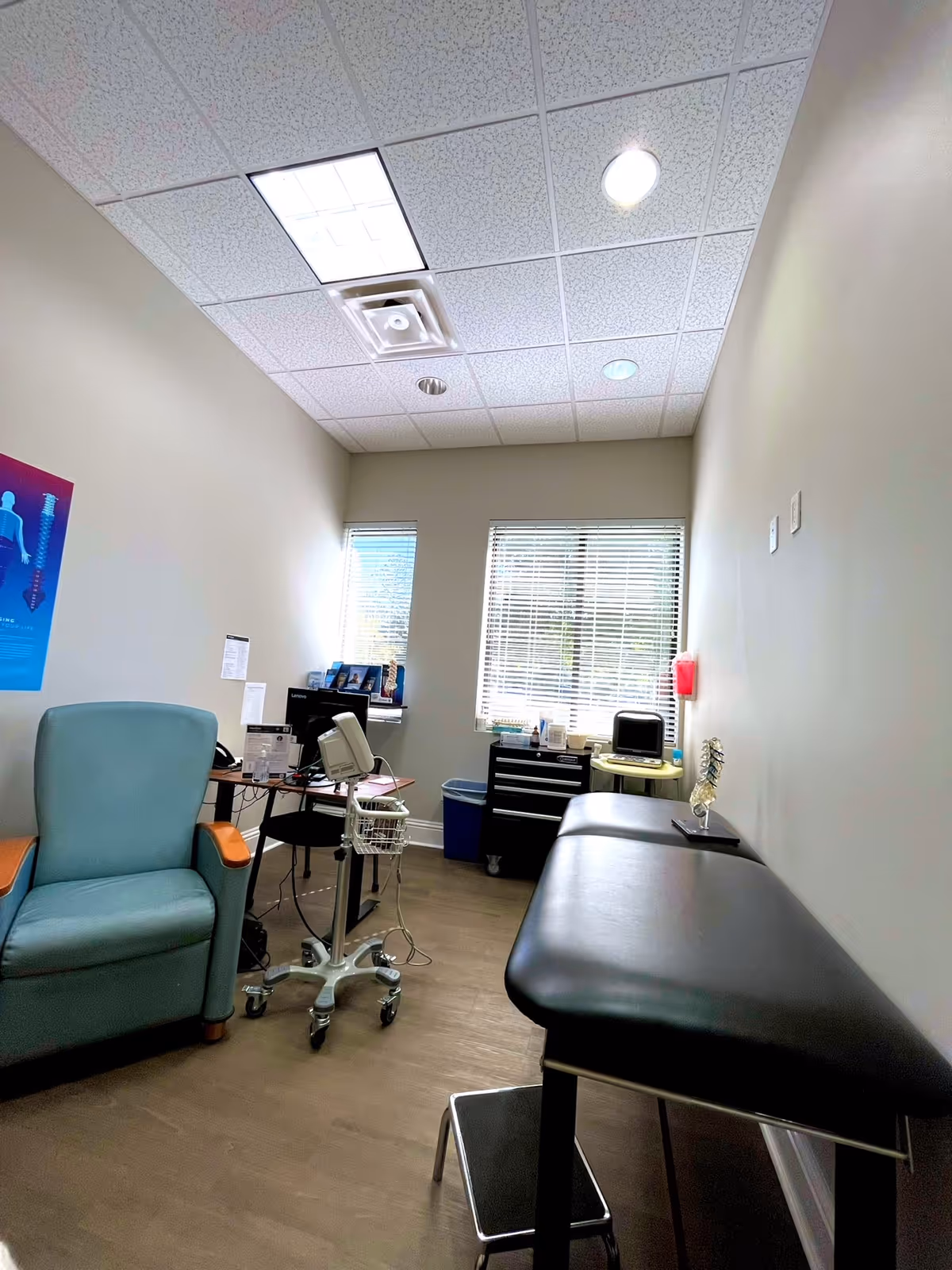 Medical examination room with teal chair, black exam table, computer, and skeleton model.