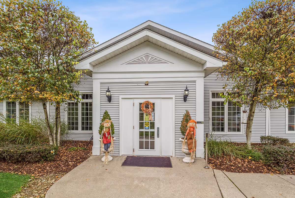 White, single-story building front with entrance, wreath, and autumn scarecrows.