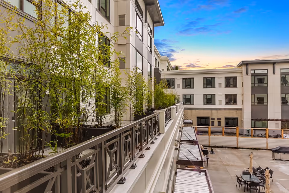 A balcony lined with bamboo planters and a decorative railing overlooking a courtyard and adjacent multi-story building at sunset.