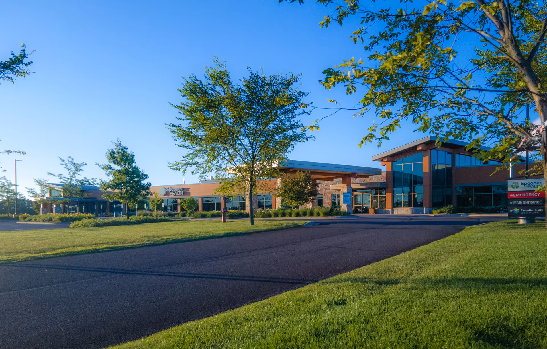 Spooner Health facility exterior with parking lot, trees, and a modern glass-front entrance.