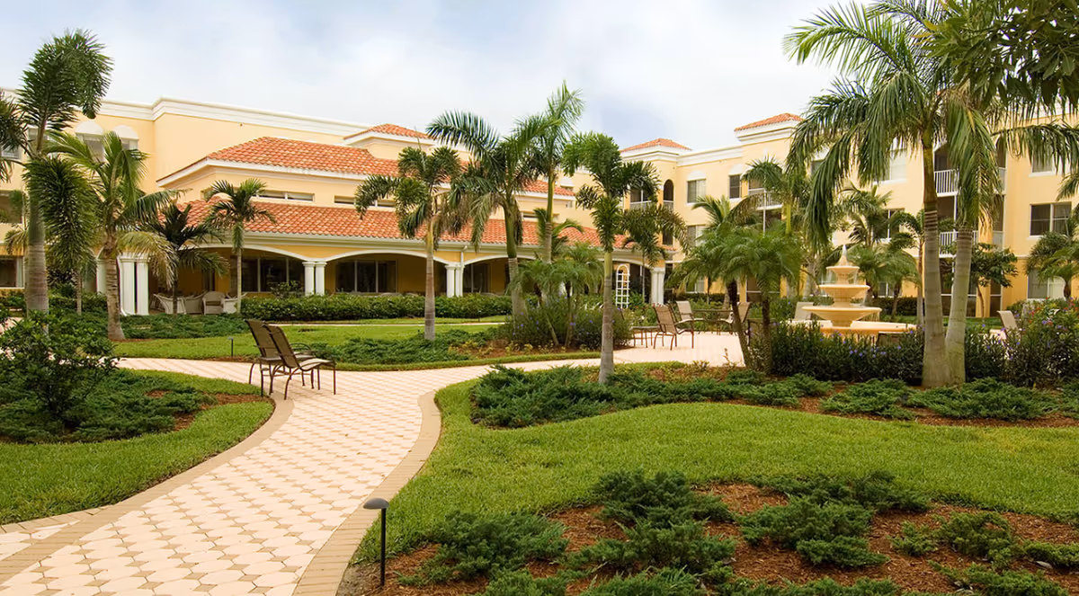 Outdoor courtyard area of The Carlisle Naples senior living facility featuring a paved walkway, palm trees, green shrubs, lawn, several chairs, and a multi-tiered water fountain in front of a beige building with red-tiled roofs.