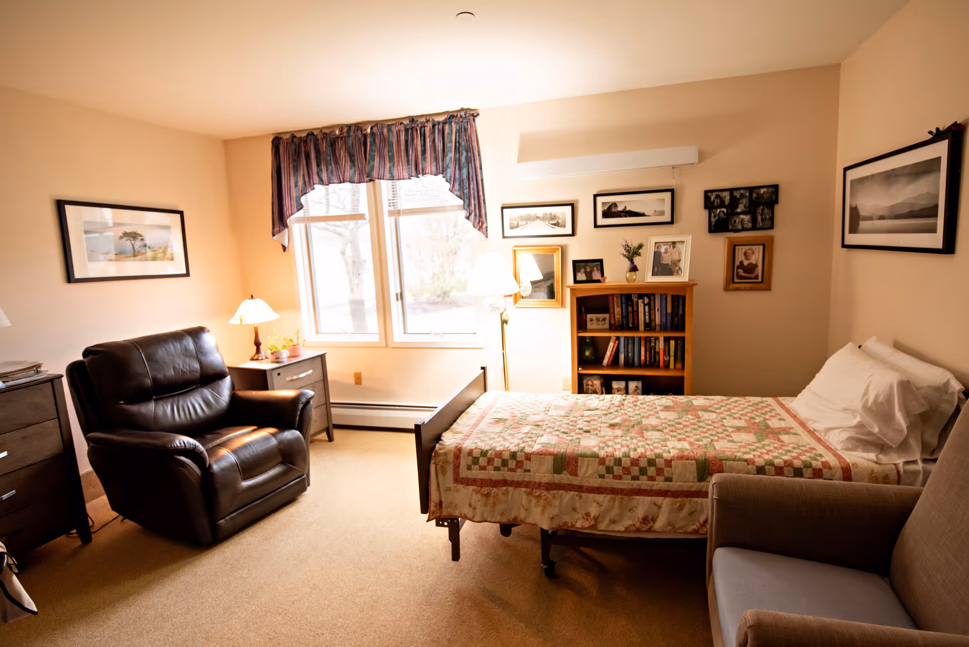 A cozy senior bedroom with a single bed covered by a patchwork quilt, armchairs, a bookshelf, and a window with curtains.