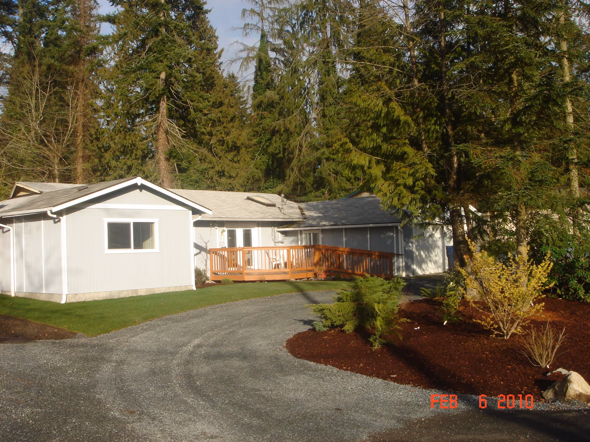 Exterior view of a single-story light gray building with a wooden ramp leading to the entrance, surrounded by green grass, mulch beds with small shrubs, and tall evergreen trees in the background under a partly cloudy sky.