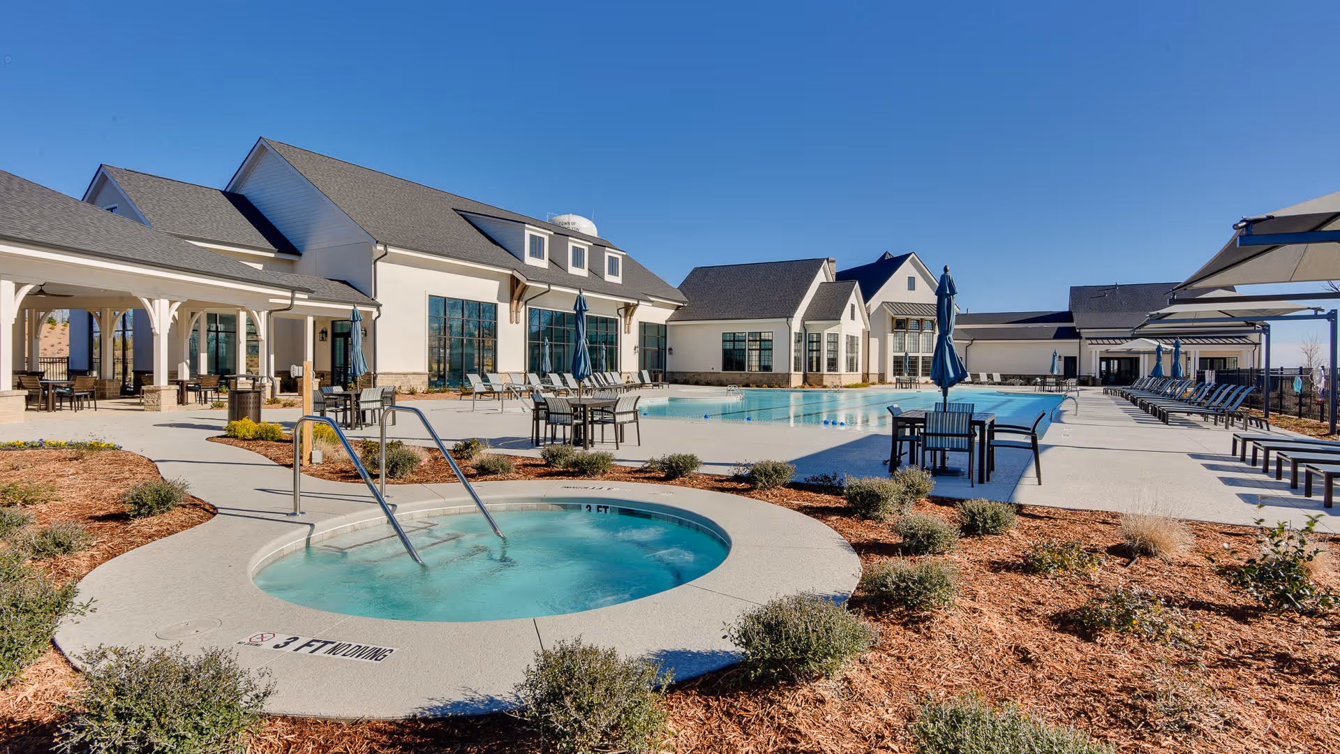 Outdoor pool area at Del Webb Chateau Elan 55+ Retirement Community featuring a hot tub in the foreground, a large swimming pool with lounge chairs and tables with umbrellas, and a clubhouse building in the background under a clear blue sky.