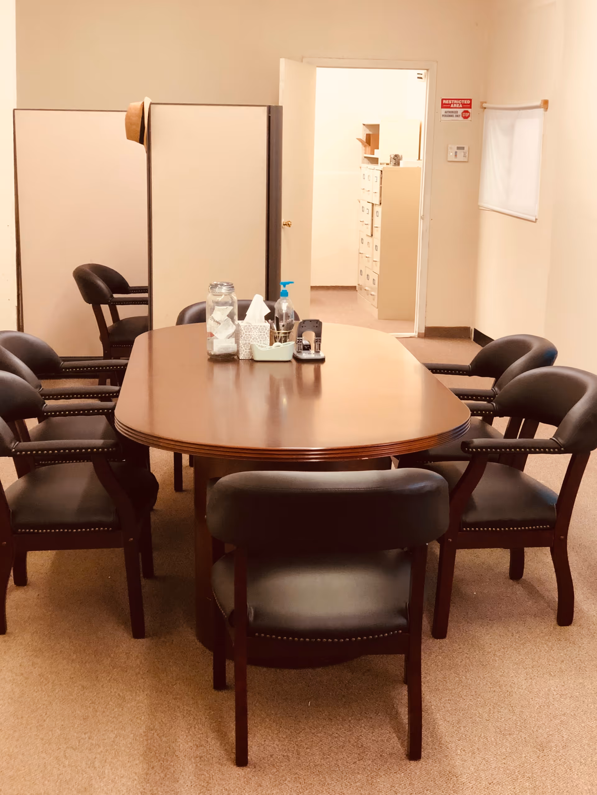 Round wooden dining table with several black chairs in a neutral, office-like room.