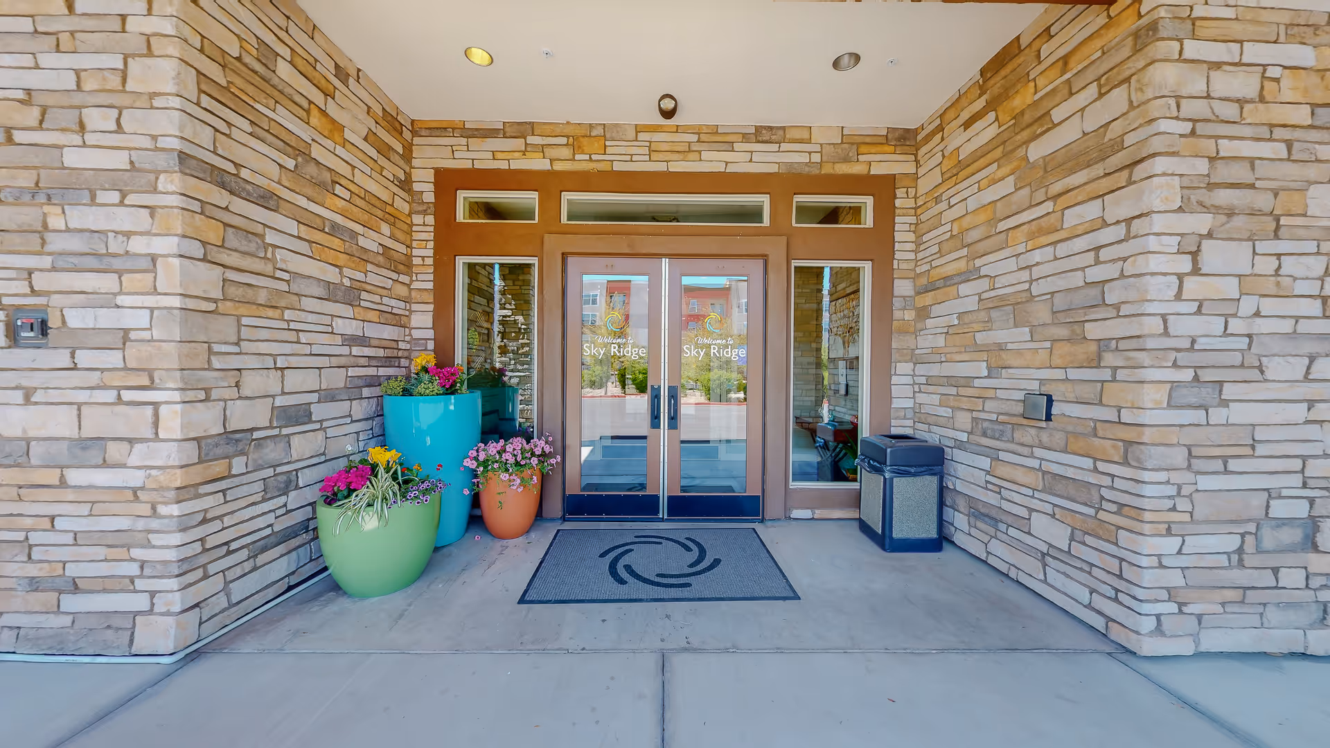 Stone-walled building entrance with glass doors, colorful planters, and a welcome mat.