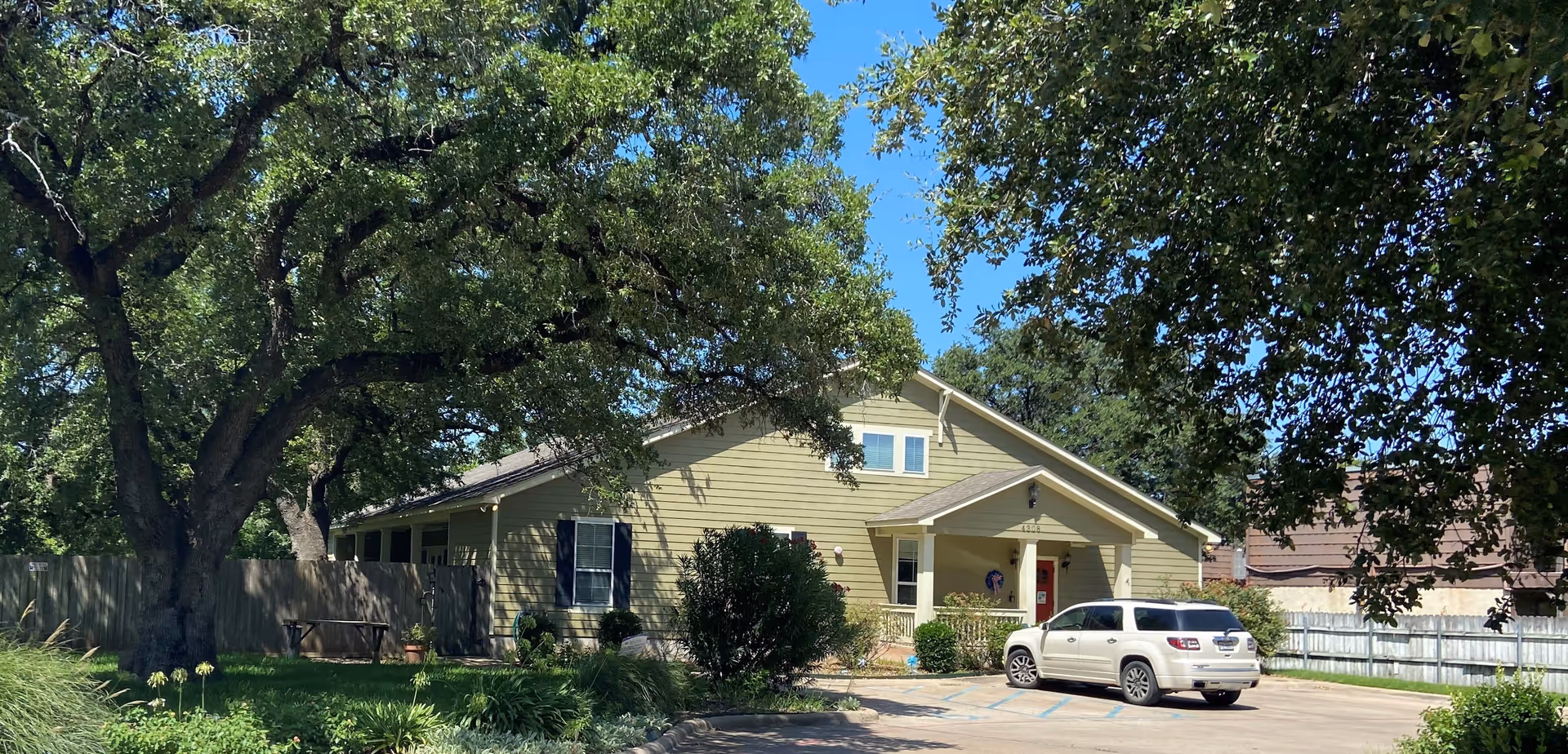 Exterior view of a single-story light yellow building with a gabled roof, surrounded by large trees and greenery. A white SUV is parked in front of the building on a paved driveway. The sky is clear and blue.